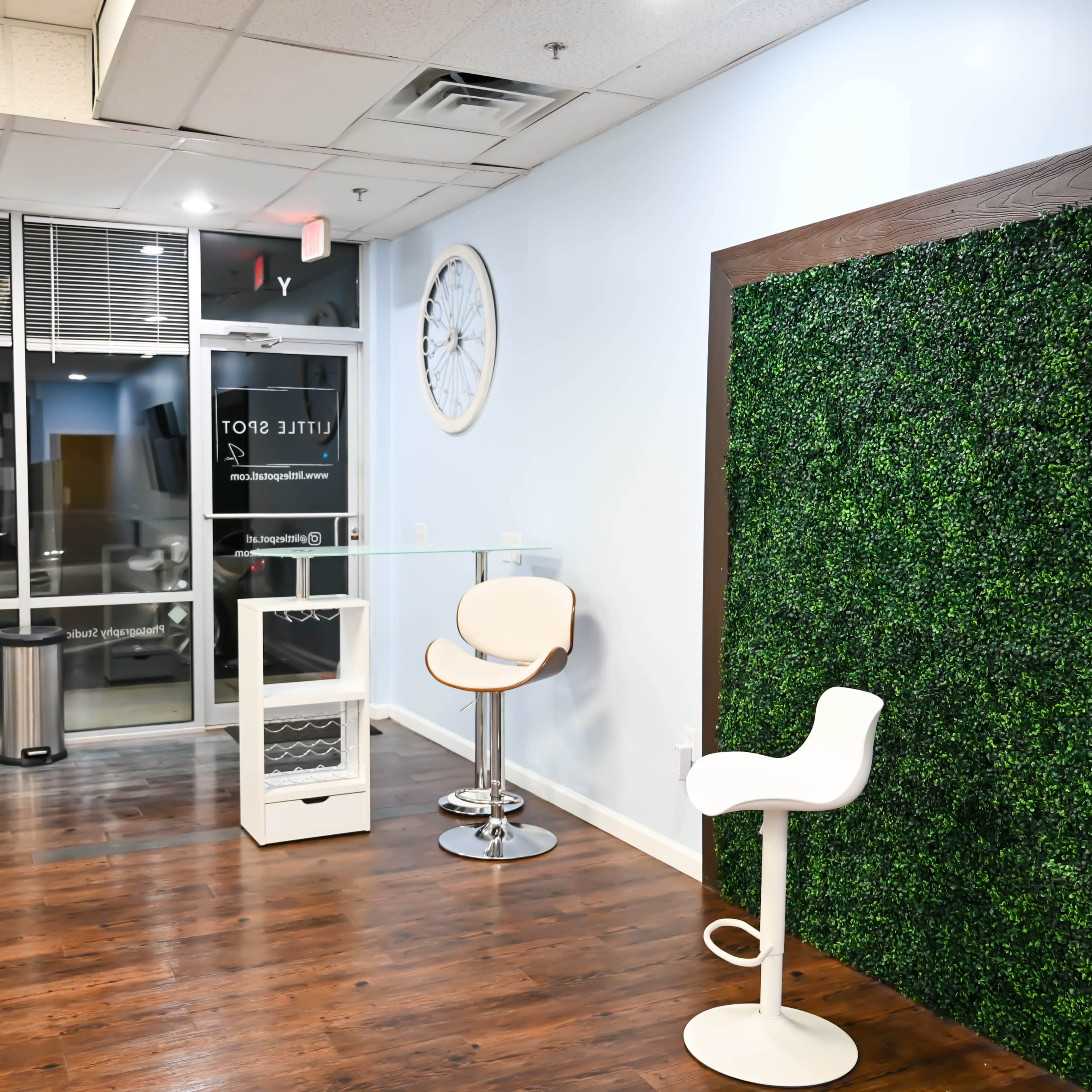 A minimalist interior space featuring a green moss wall, a glass table with two stools, and large windows with light coming in.