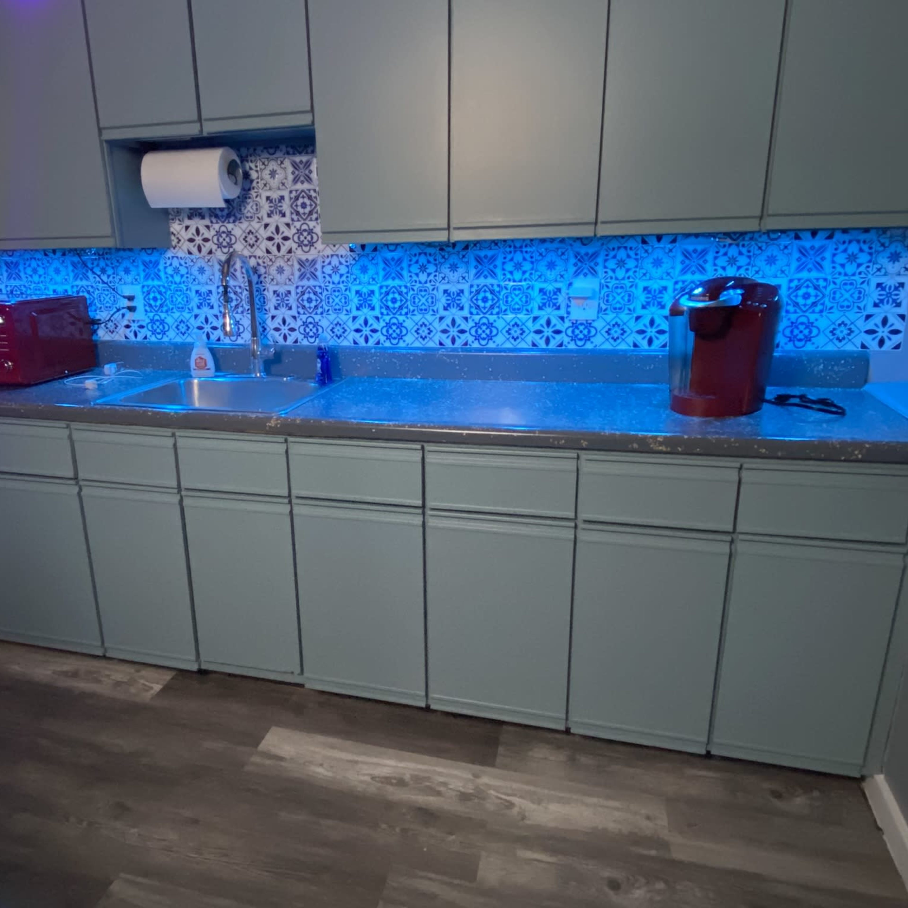 A modern kitchen with green cabinets, a countertop, a sink, a microwave, and a blue-tiled backsplash illuminated by blue lighting.
