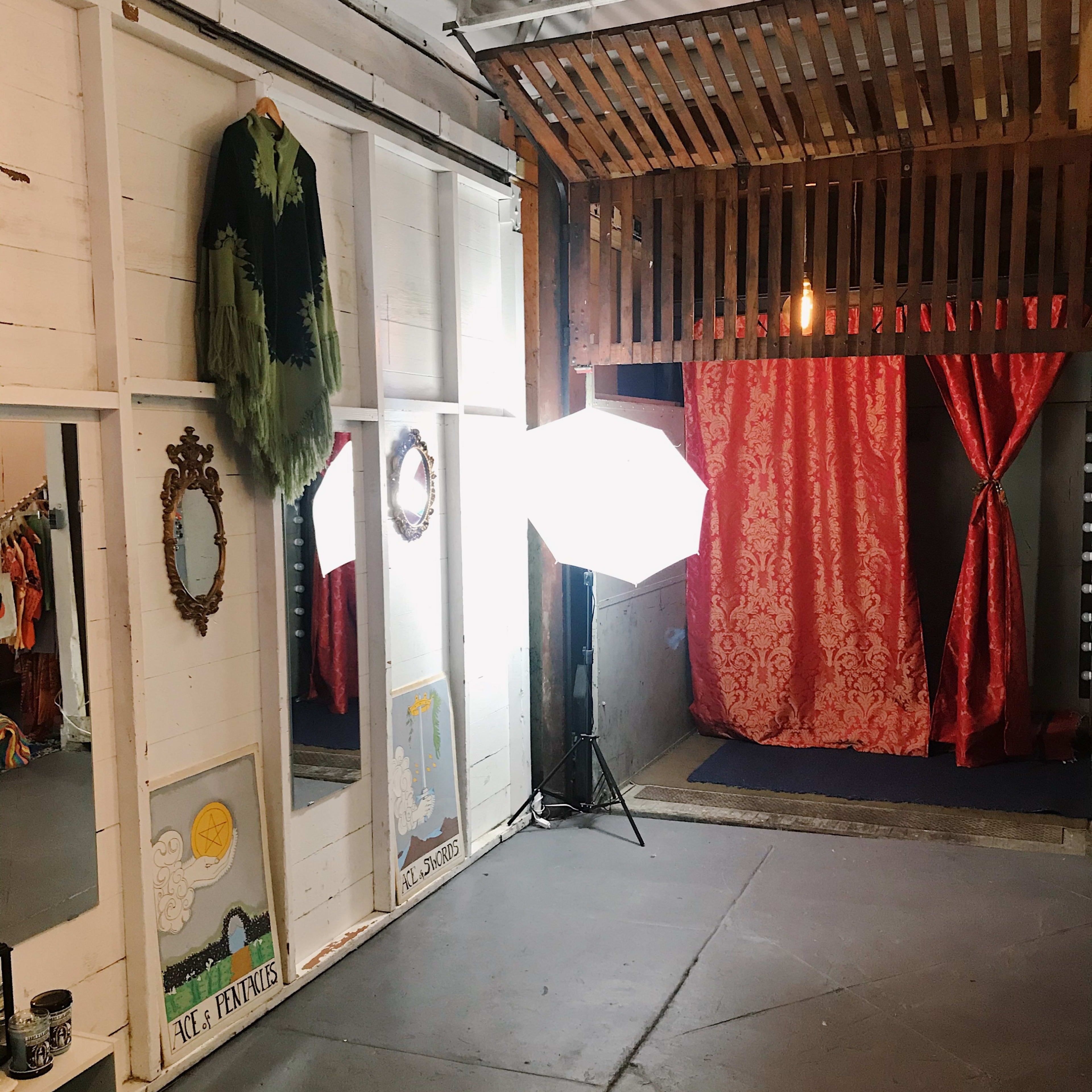 A room with mirrors, a garment hanging on the wall, and a backdrop of red curtains, illuminated by a softbox light.