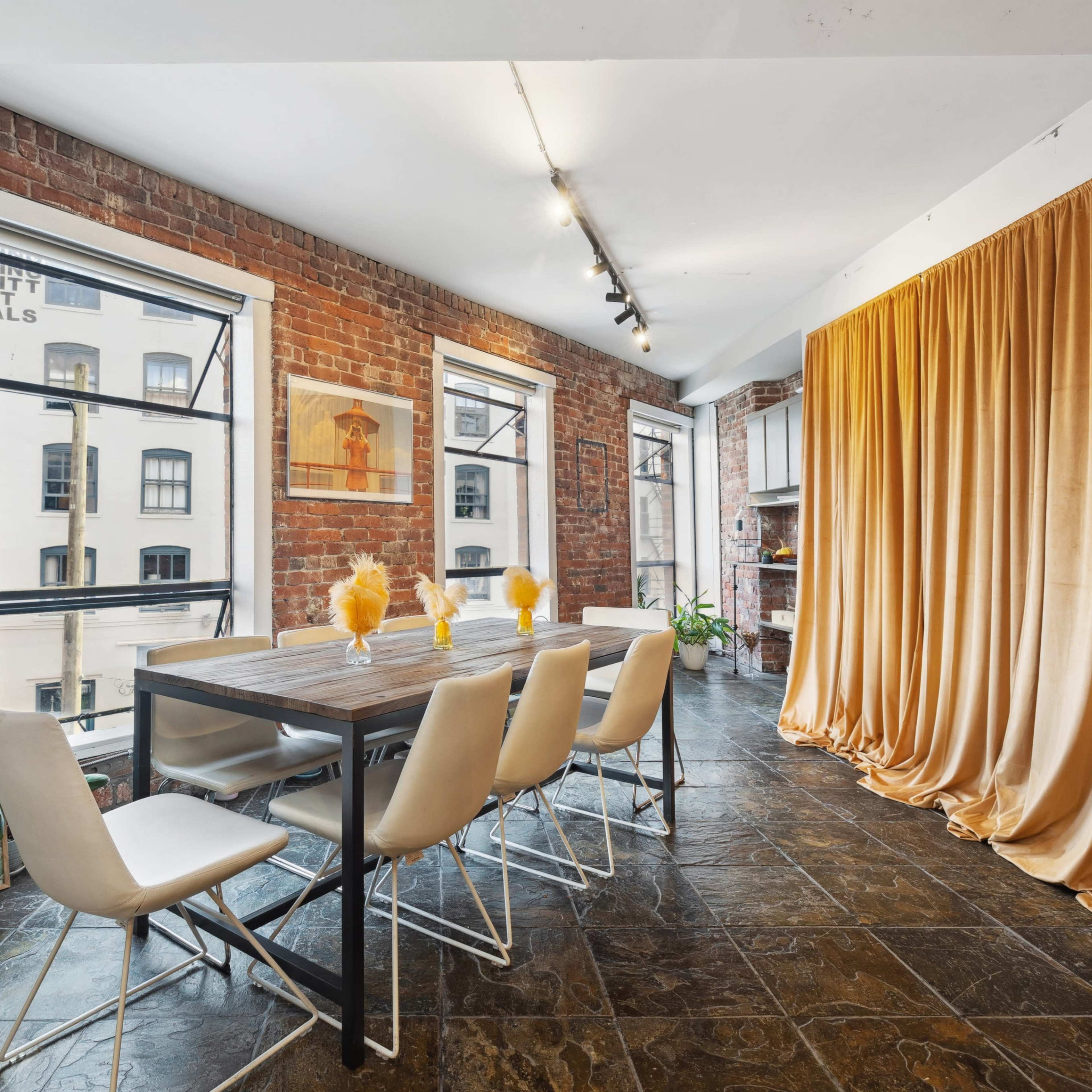 A dining area with a wooden table and six white chairs, surrounded by exposed brick walls and large windows, with a yellow curtain draped in the corner.