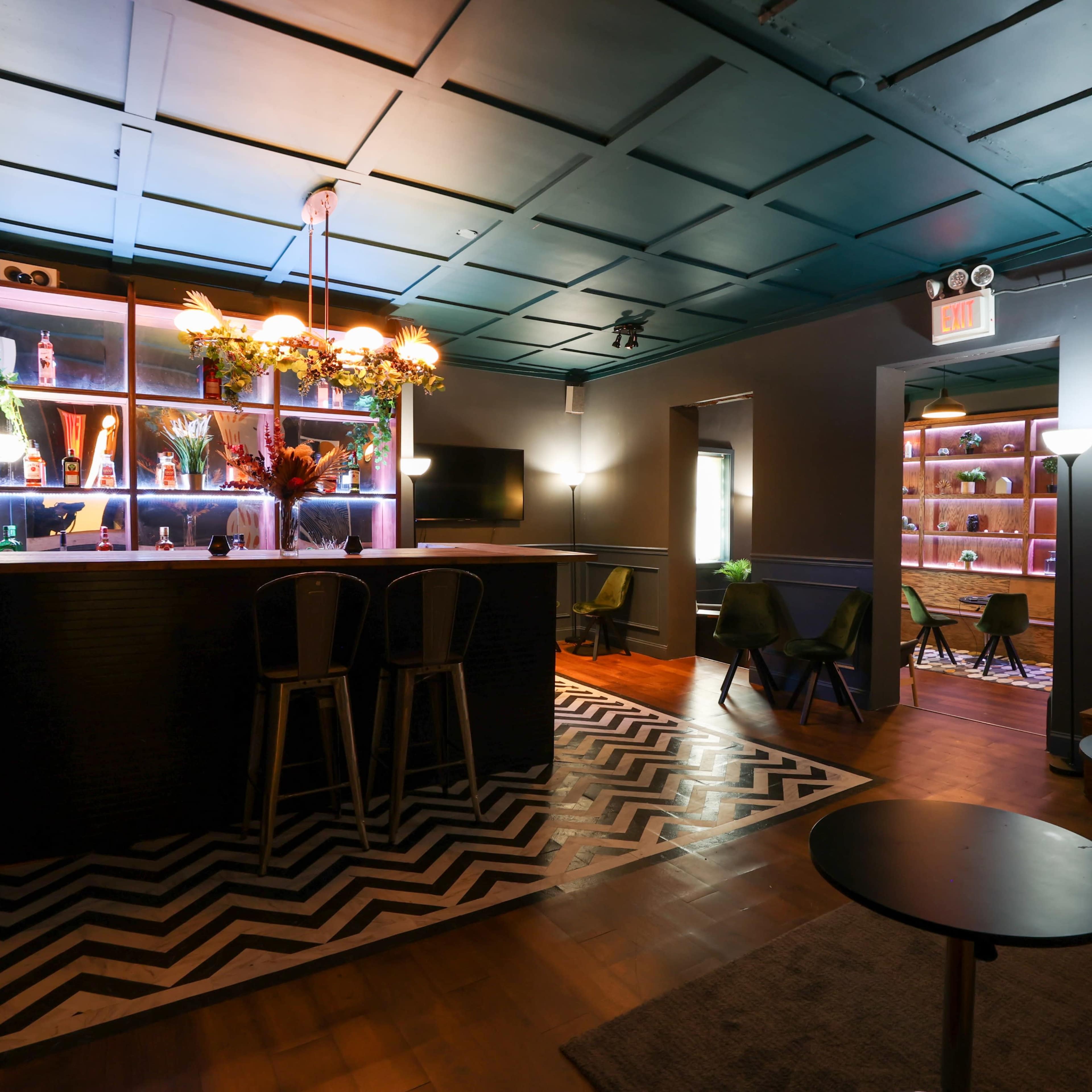 A modern lounge area featuring a bar with high stools, decorative shelving, and a patterned rug on wooden flooring.