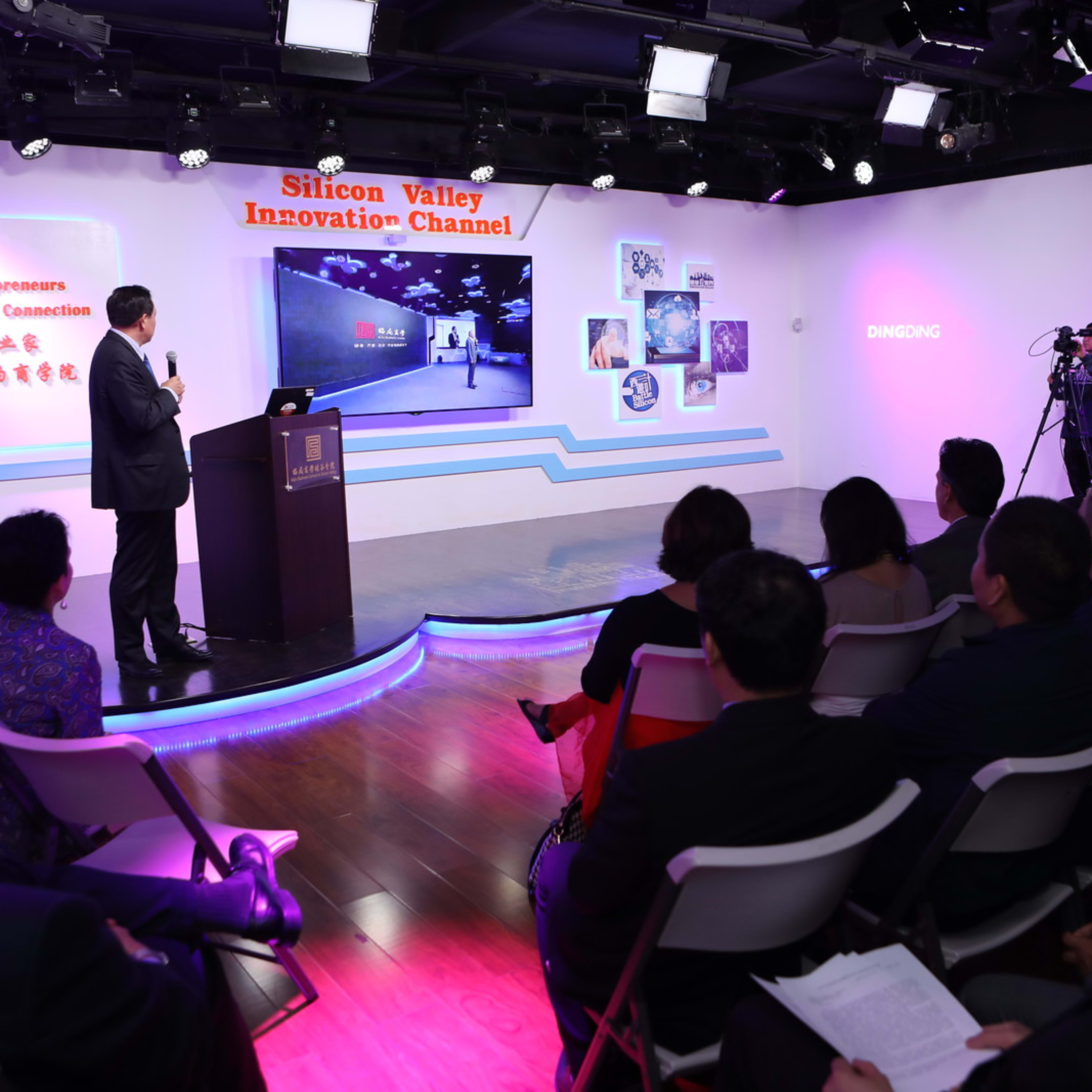 A speaker presents in front of an audience at the Silicon Valley Innovation Channel event, with cameras capturing the presentation on a screen.