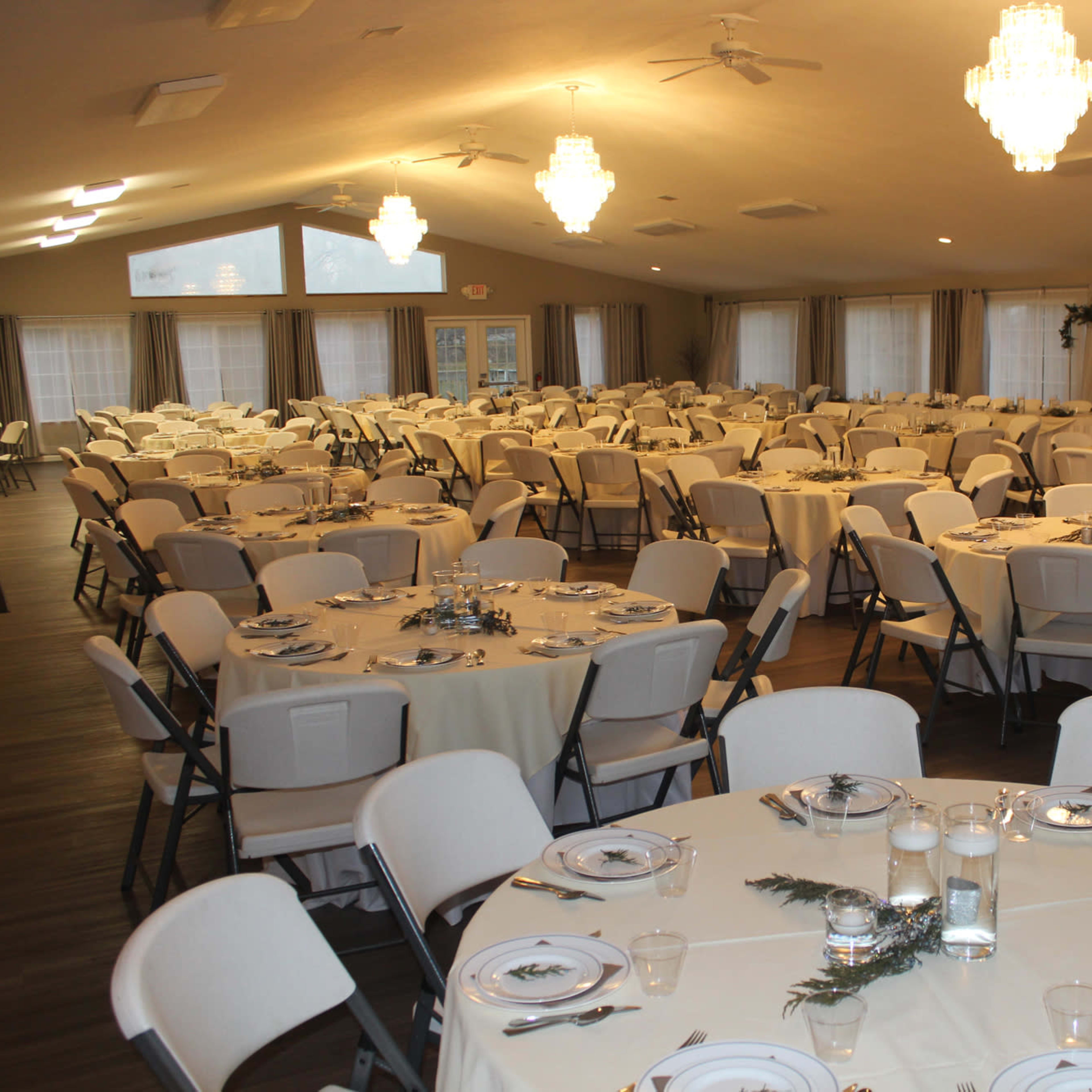 A large banquet hall set up with numerous round tables, each covered with white tablecloths and arranged with plates and glassware.