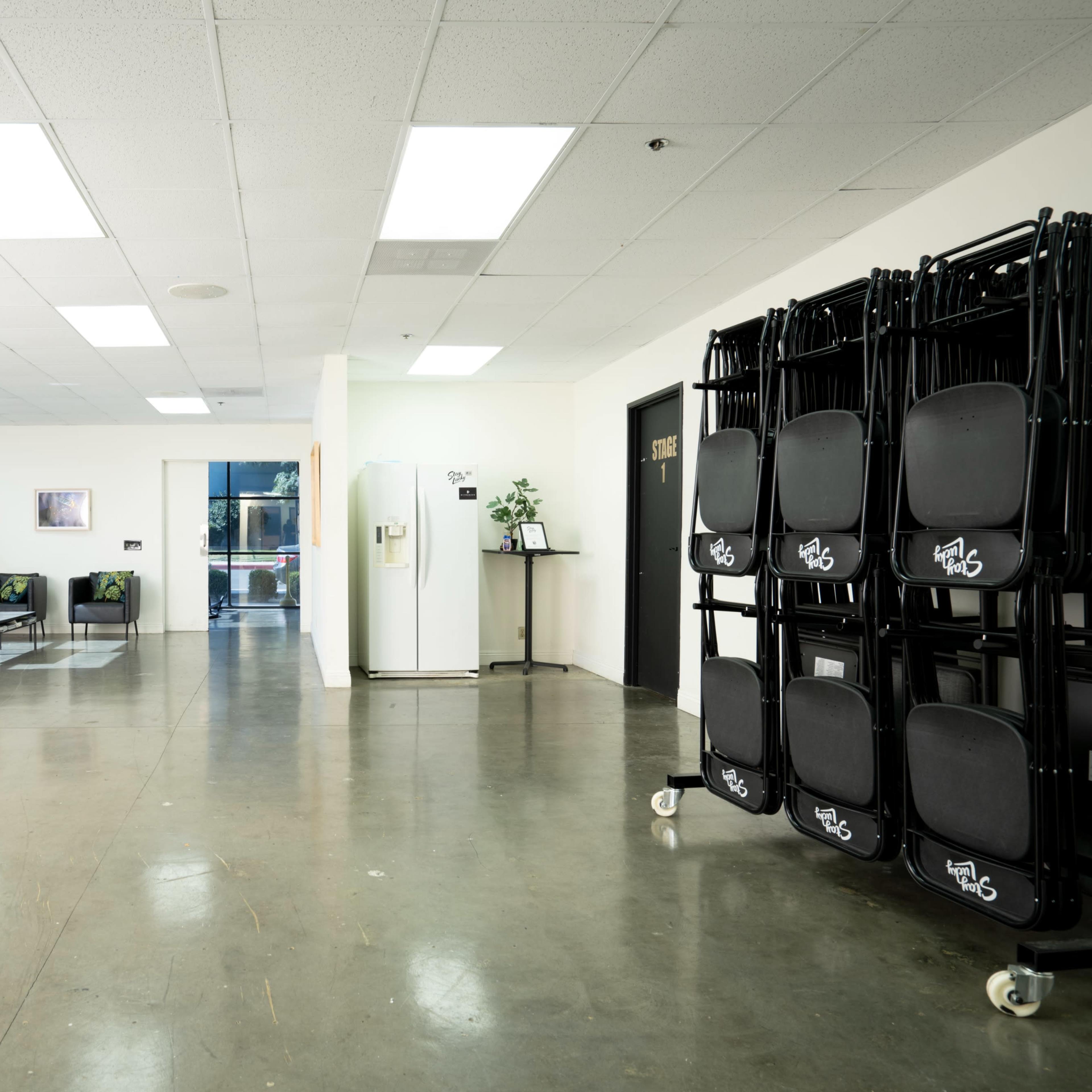 A spacious, well-lit interior with a seating area, a refrigerator, and stacked chairs against the wall.