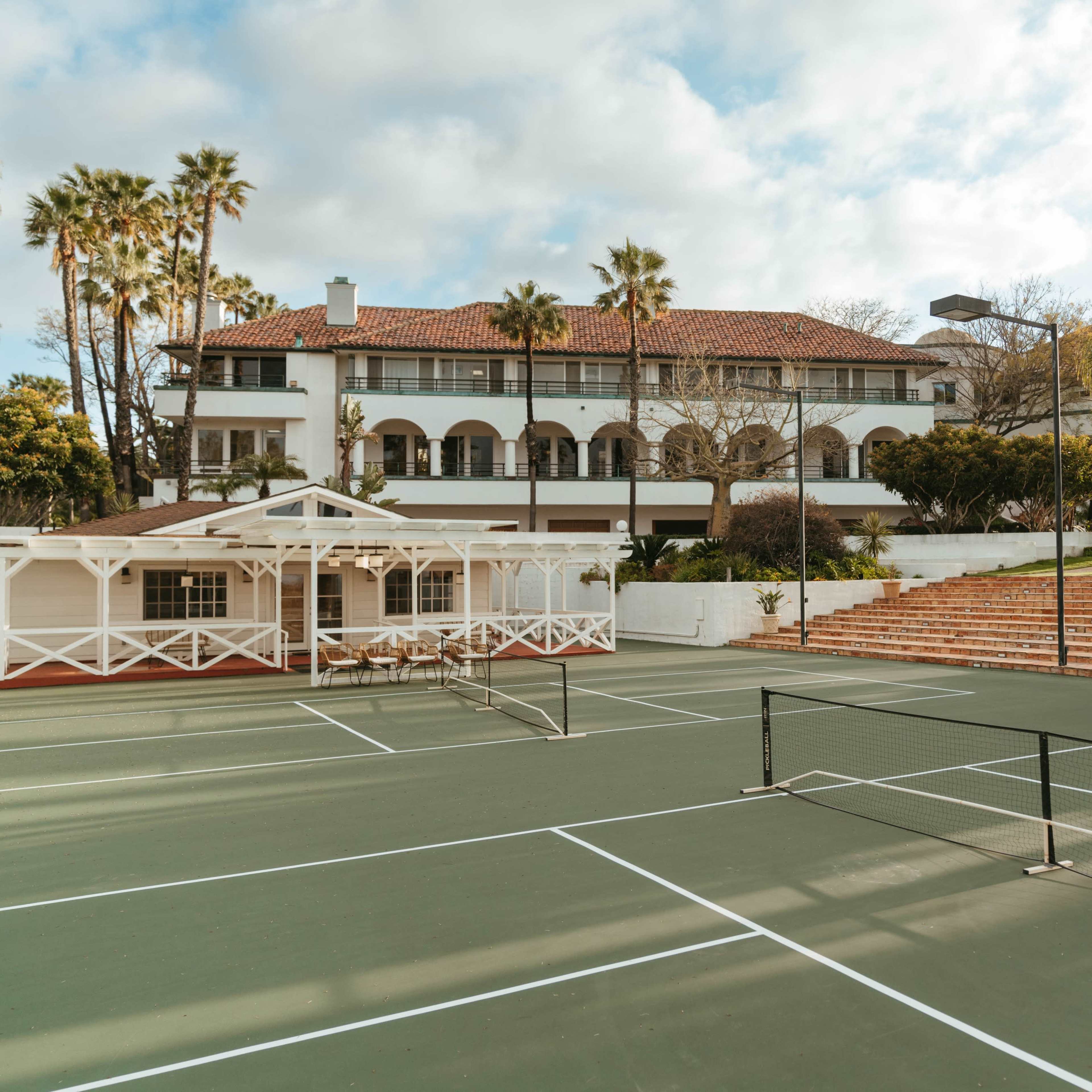 A tennis court with two nets and a large white building featuring balconies and palm trees in the background.