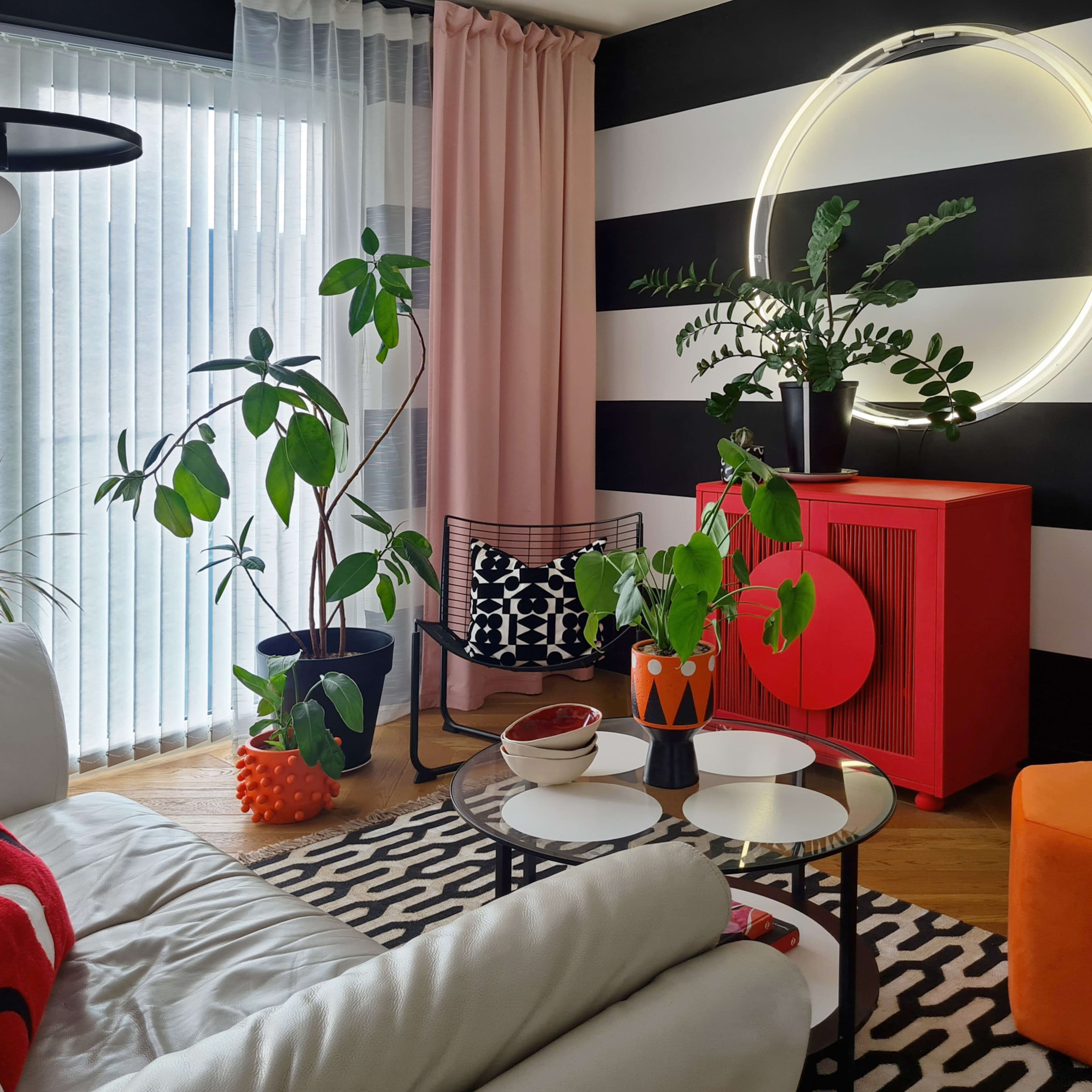 The living room features a white leather sofa, a round glass coffee table, and various indoor plants, set against a backdrop of black and white striped walls and a red cabinet.