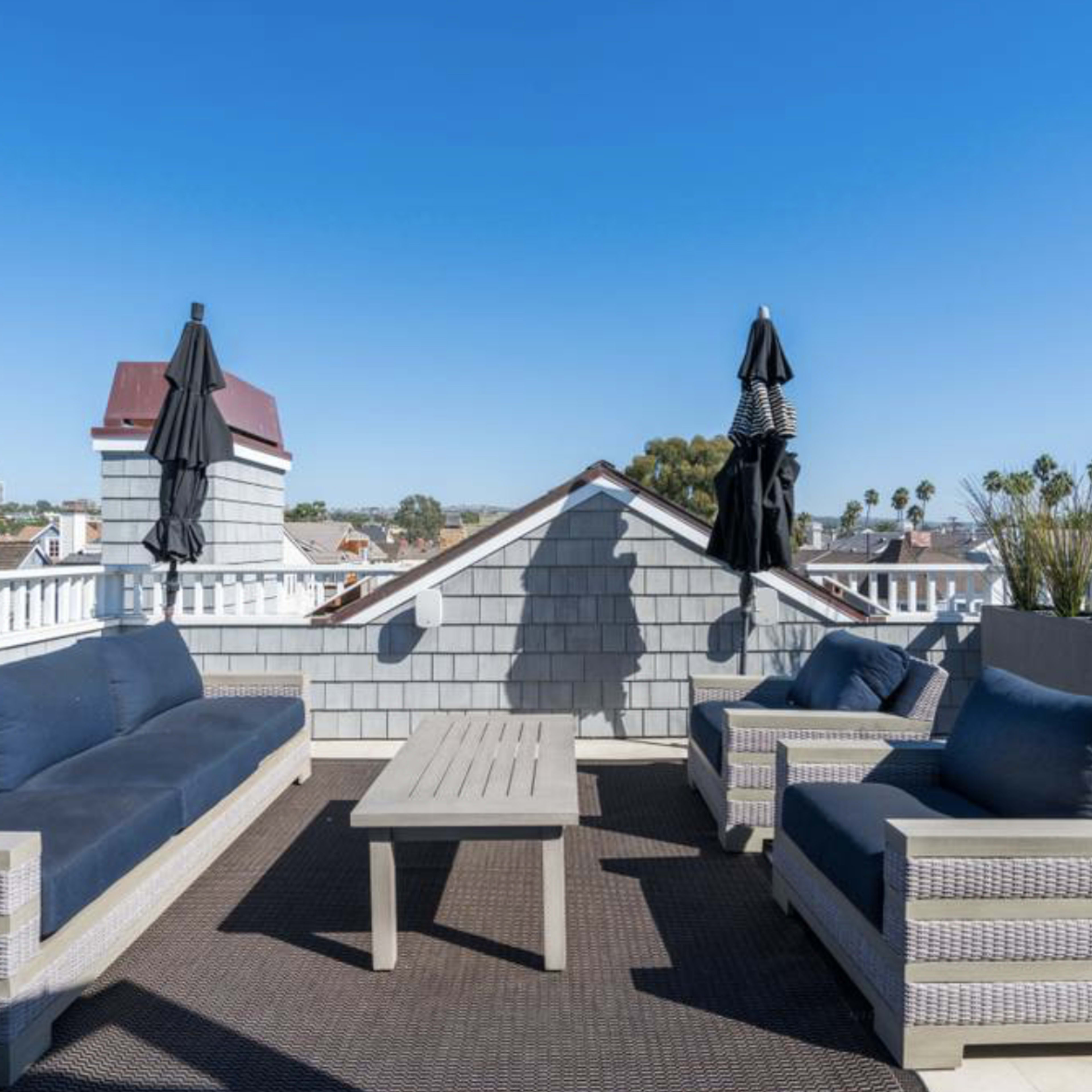A rooftop terrace furnished with two sofas, a coffee table, and two umbrellas, overlooking a view of the surroundings featuring palm trees and clear blue skies.