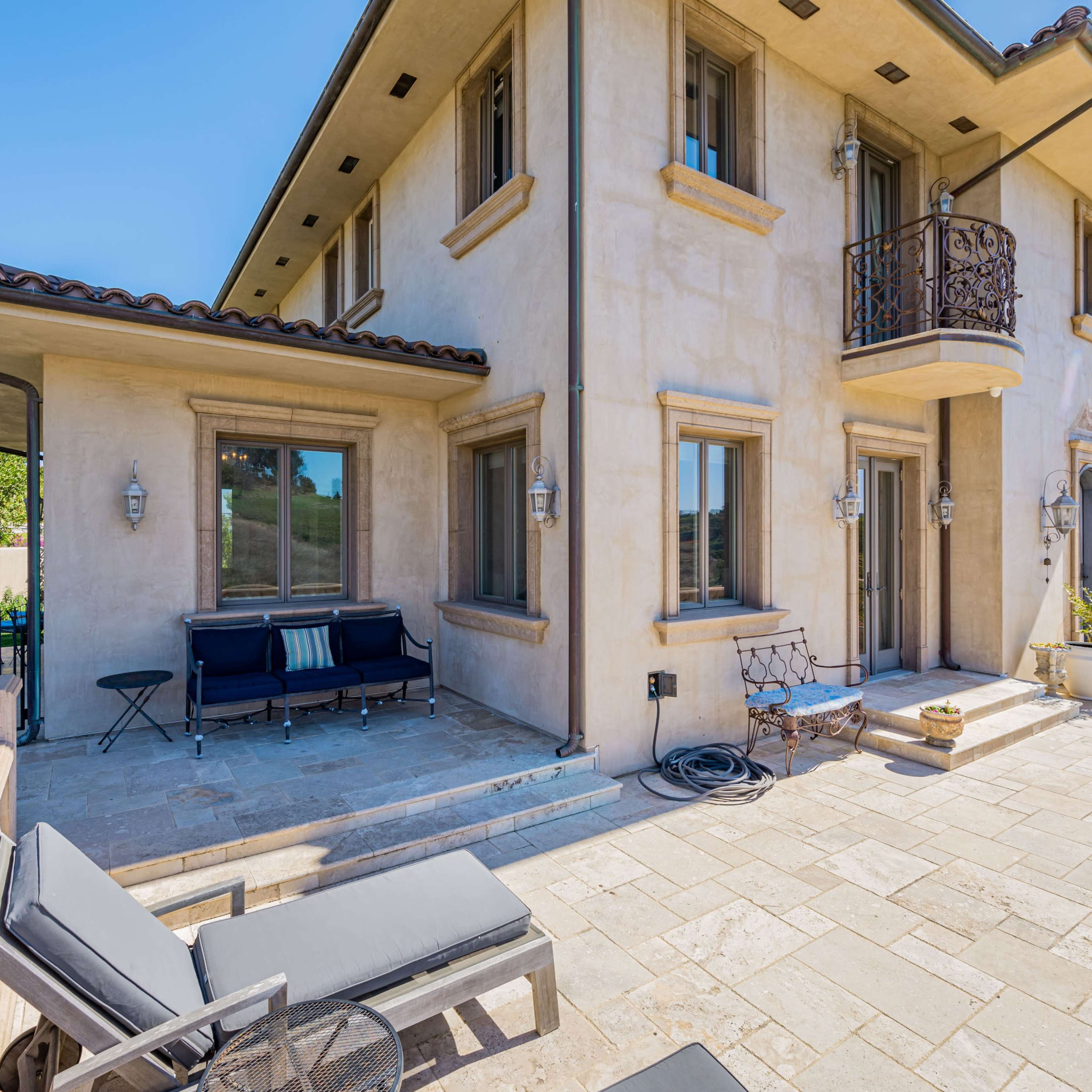 A stucco house with a balcony overlooking a patio area, featuring lounge chairs and potted plants.