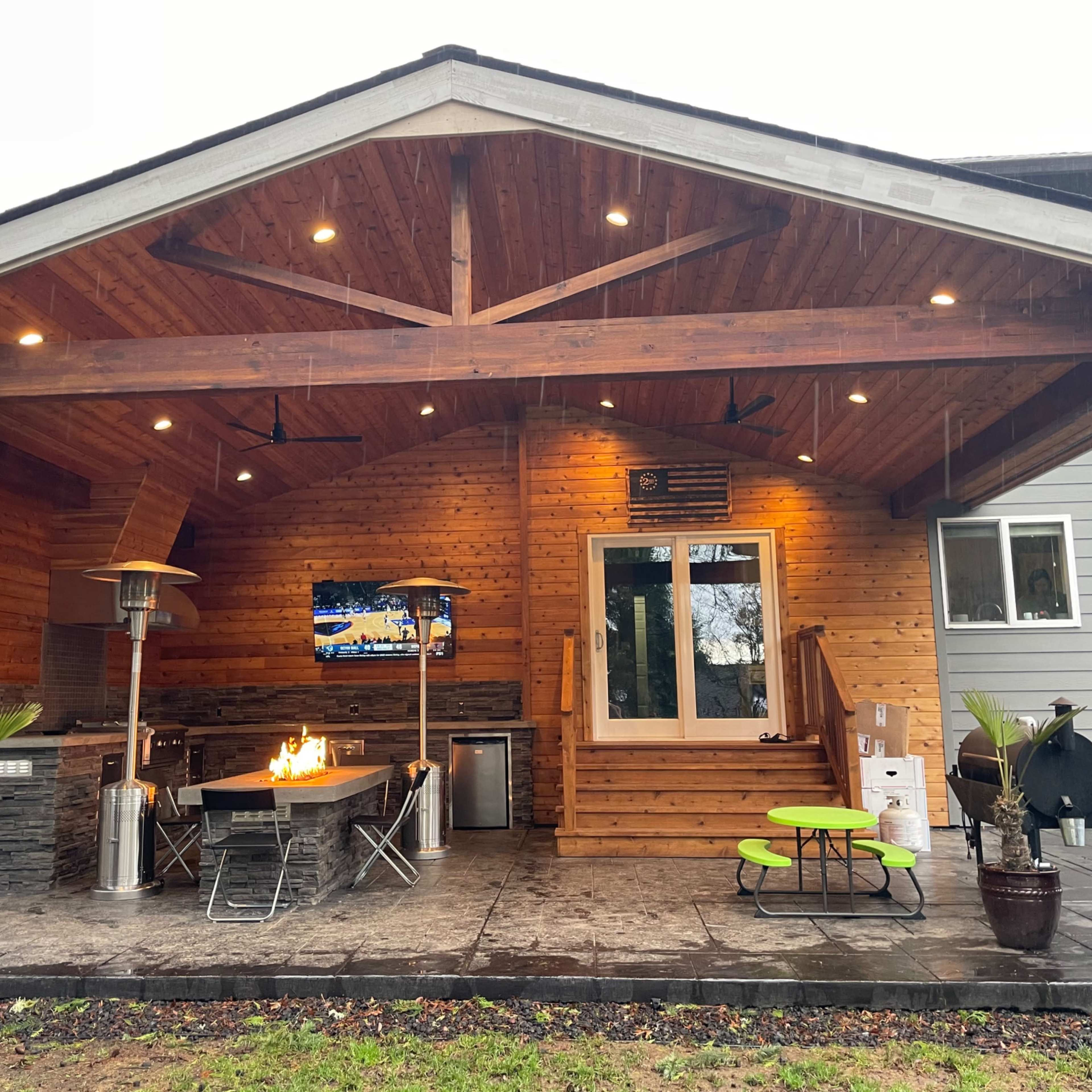 A covered outdoor patio featuring a grill, a dining table with chairs, a fire pit, and a television mounted on the wall, set against a wooden backdrop.