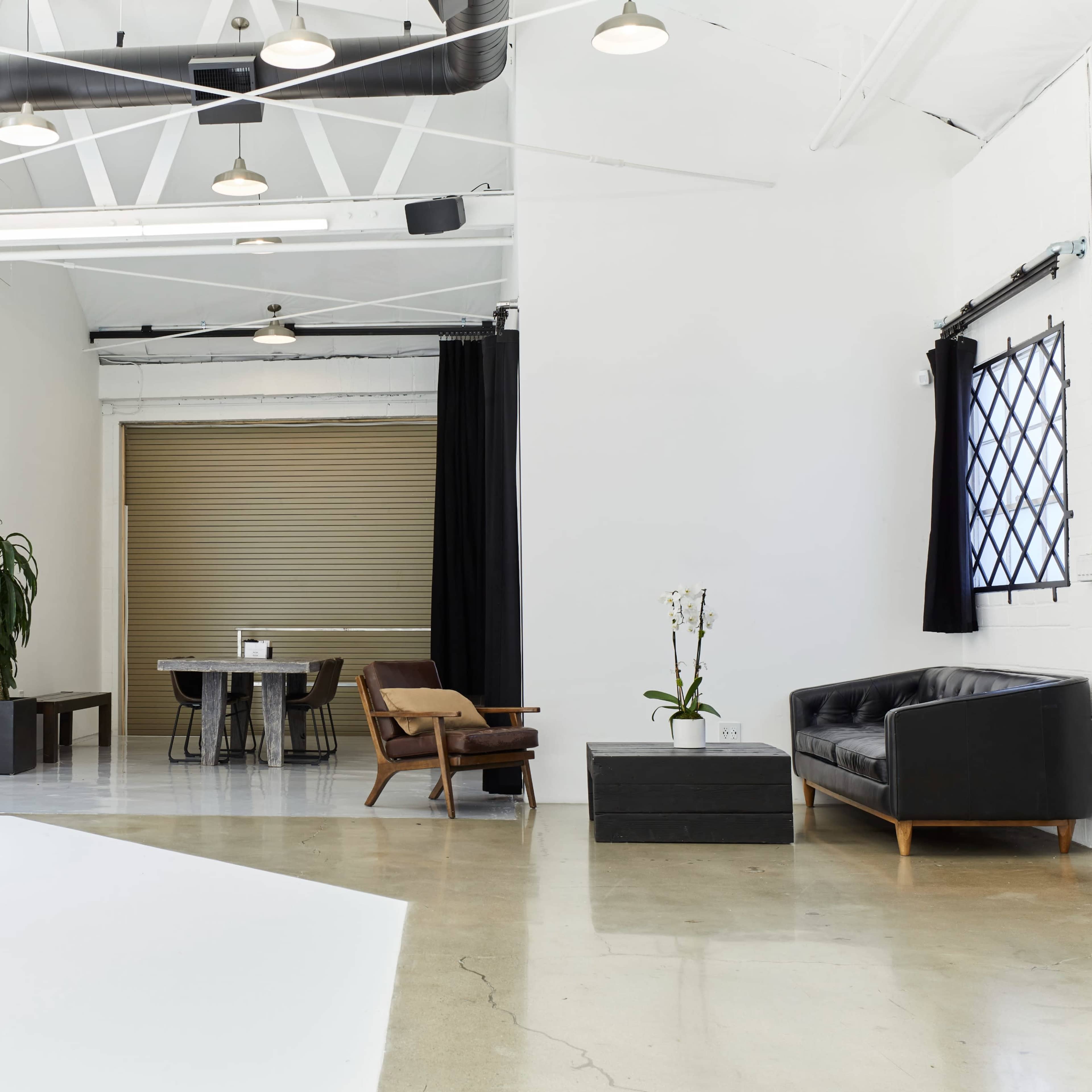 A spacious, modern interior features a black leather sofa, a wooden dining table, and large windows with dark curtains.