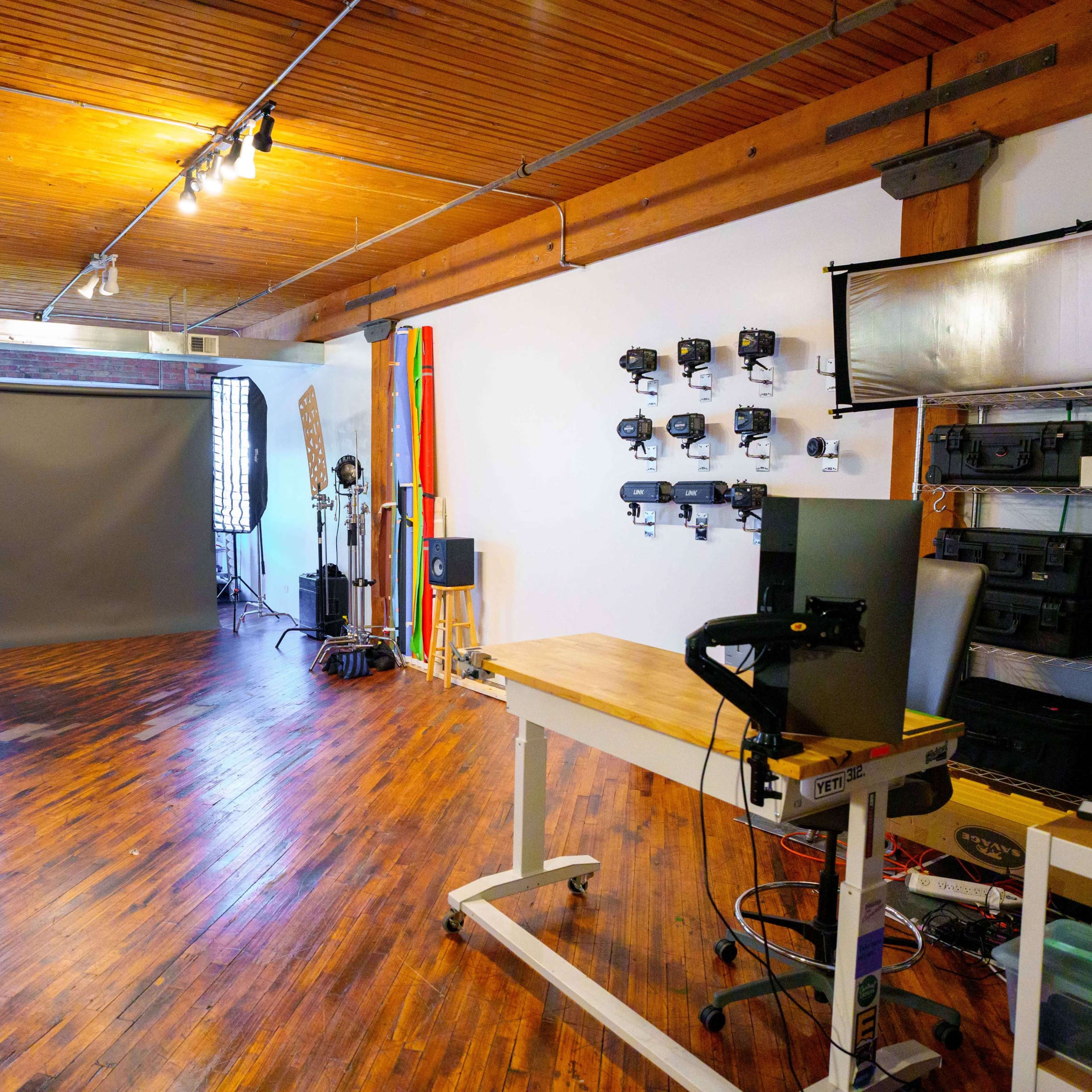 A spacious photography studio featuring a gray backdrop, professional lighting equipment, and wooden flooring.