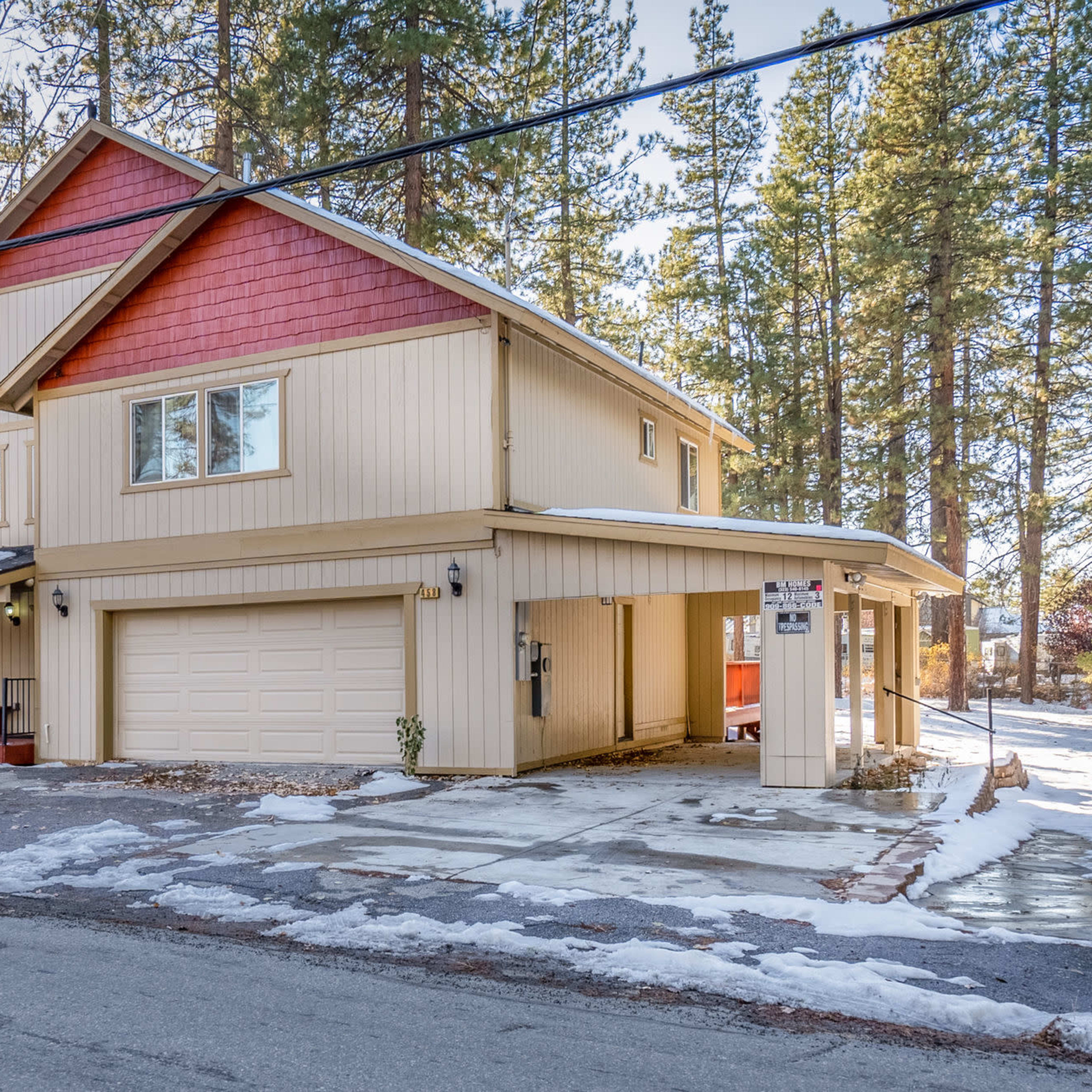A two-story house with a light-colored exterior and red accents, situated on a snowy street lined with tall pine trees.