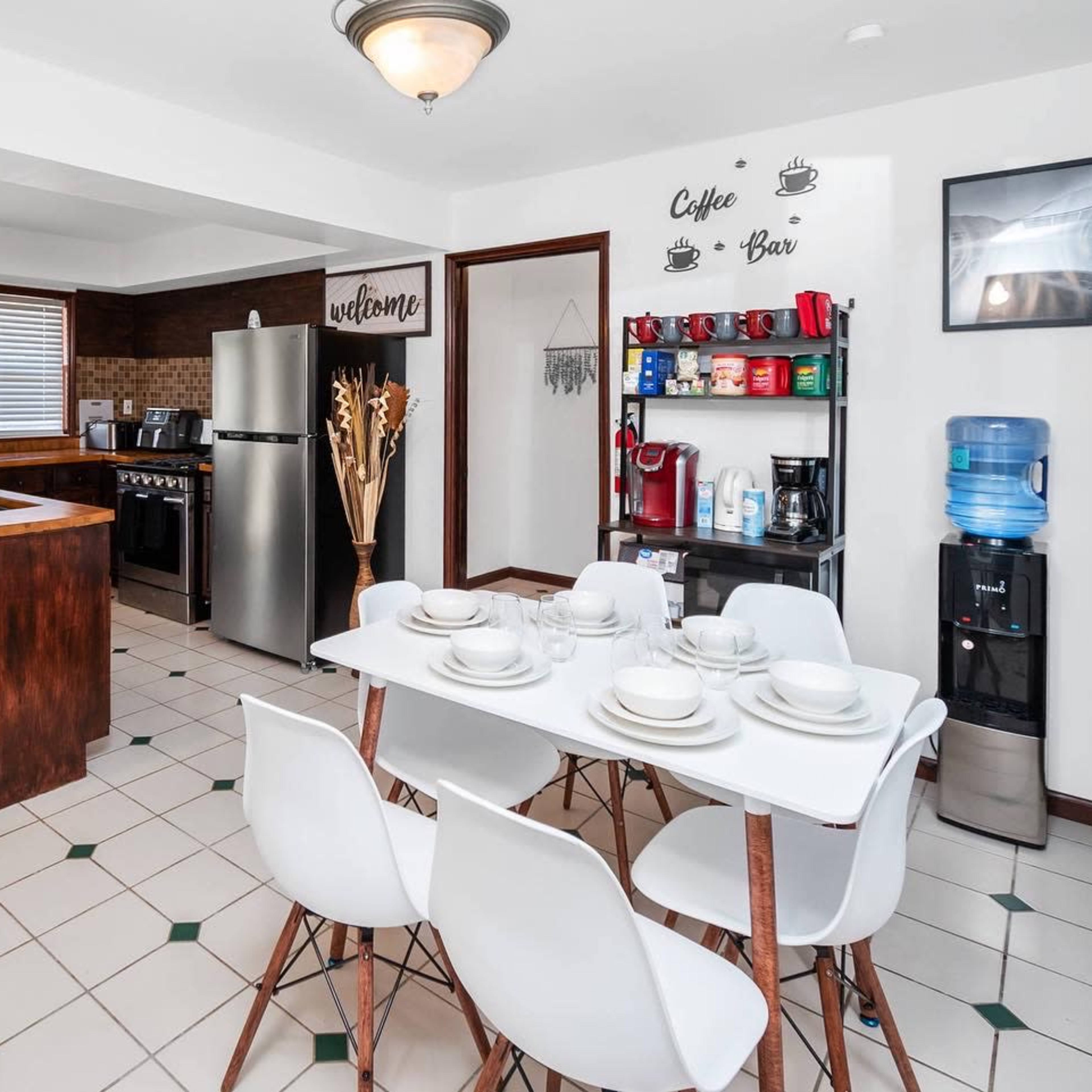 A modern kitchen with a dining table set for four, a stainless steel refrigerator, and a coffee bar along the wall.