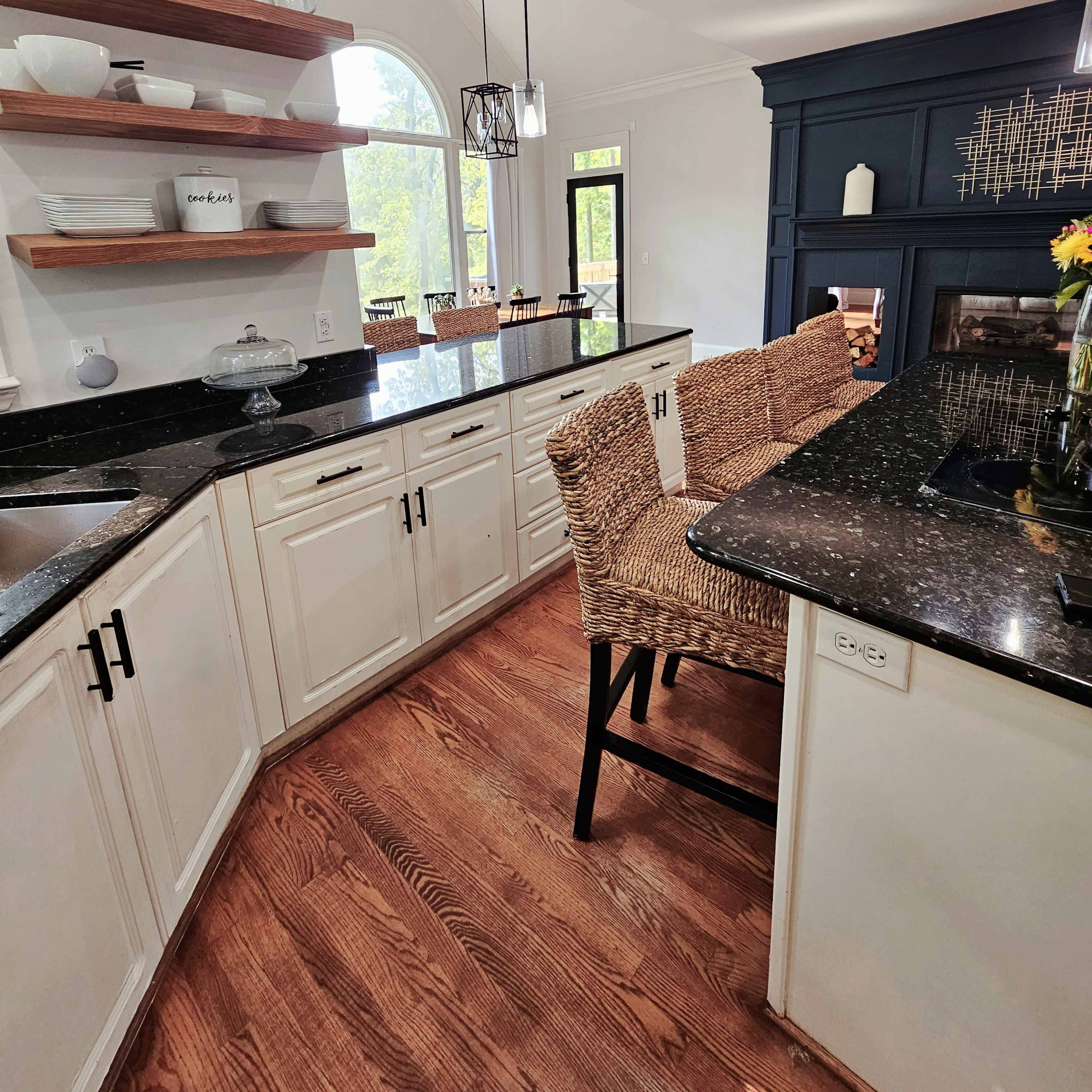 A modern kitchen featuring white cabinetry, dark granite countertops, wooden shelves, and bar stools with a view into an adjacent dining area.