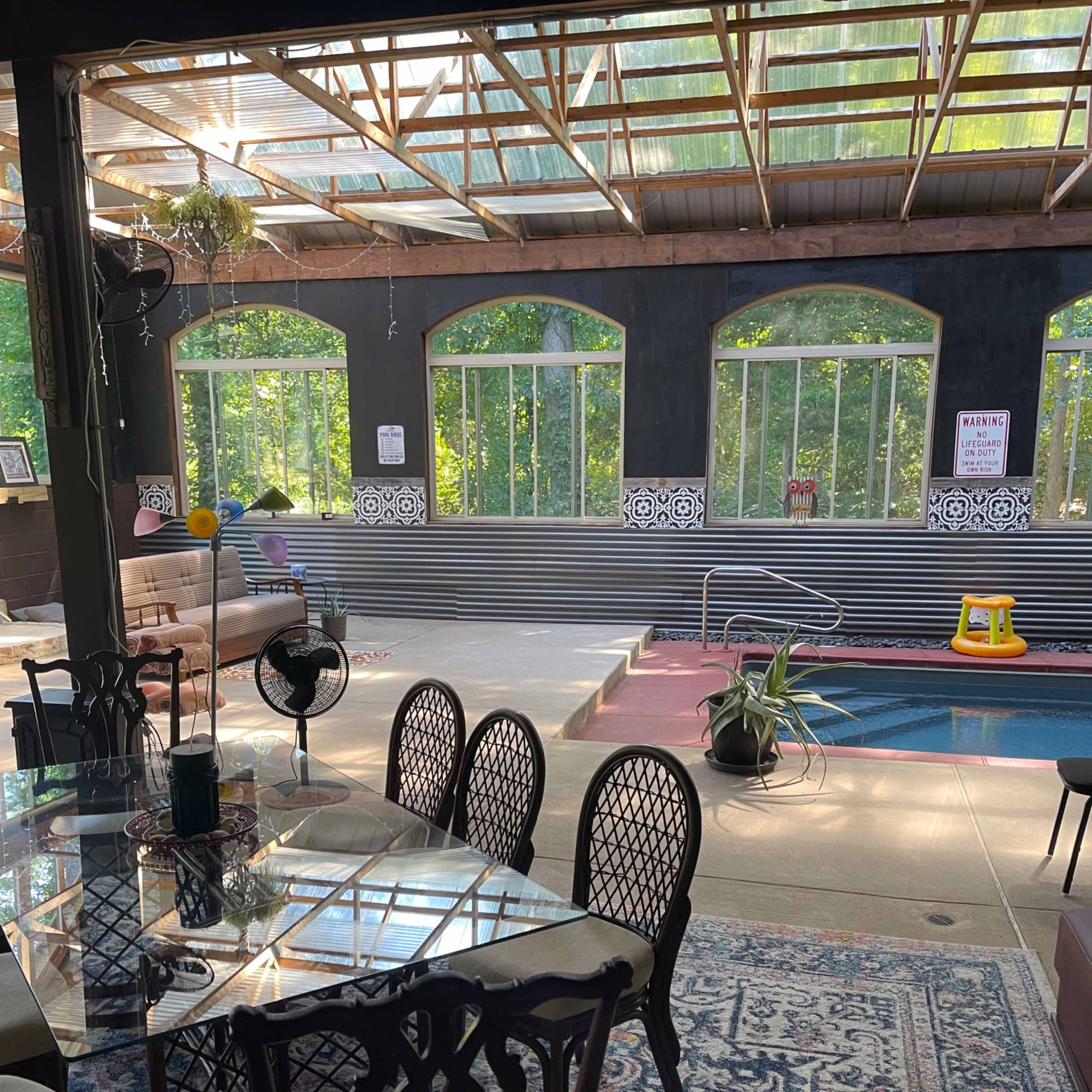 A sunlit indoor space featuring a dining area with a glass table and chairs, a swimming pool, and large windows overlooking greenery.