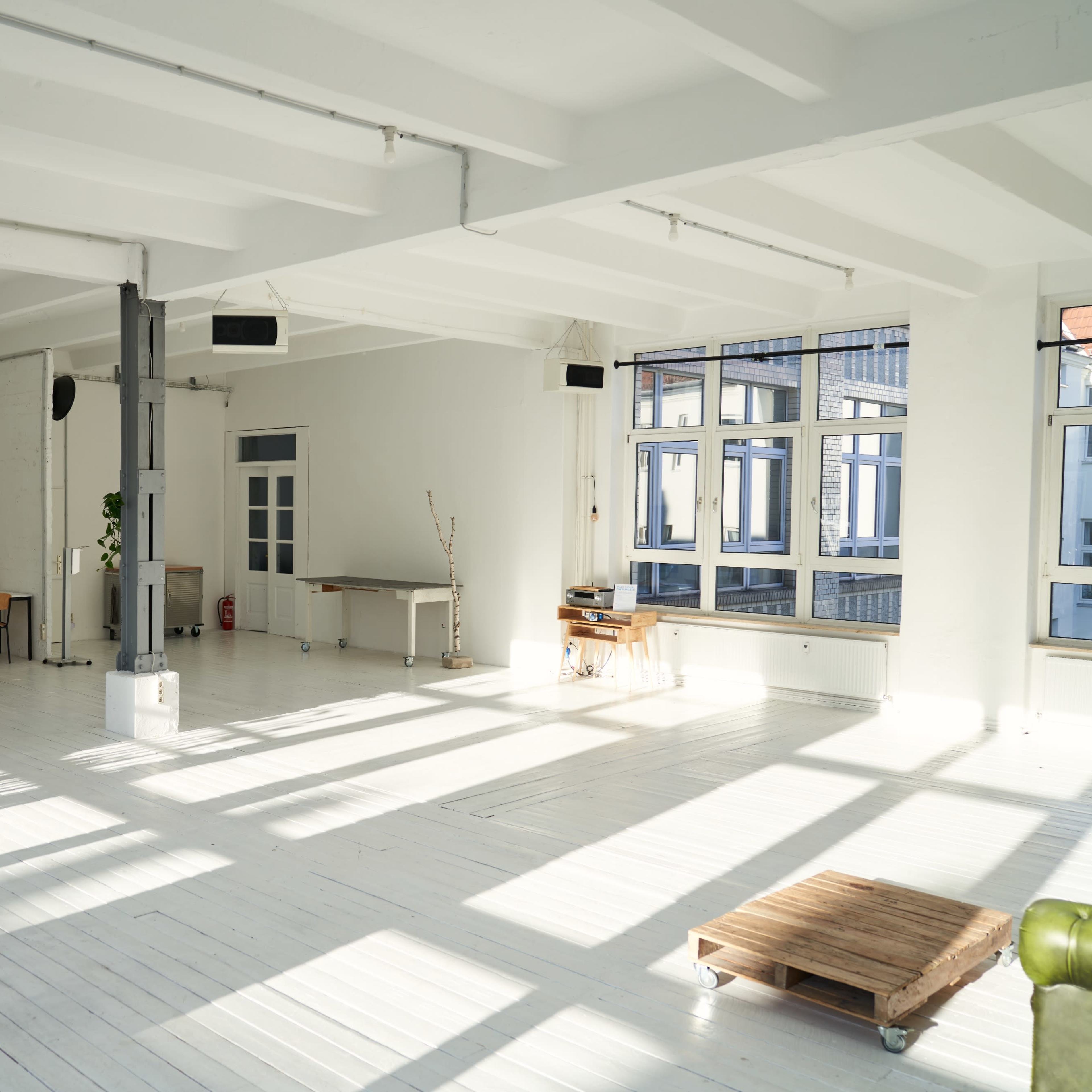 A spacious, well-lit room with white wooden floors, featuring a few tables and chairs, large windows, and shadows cast on the floor.