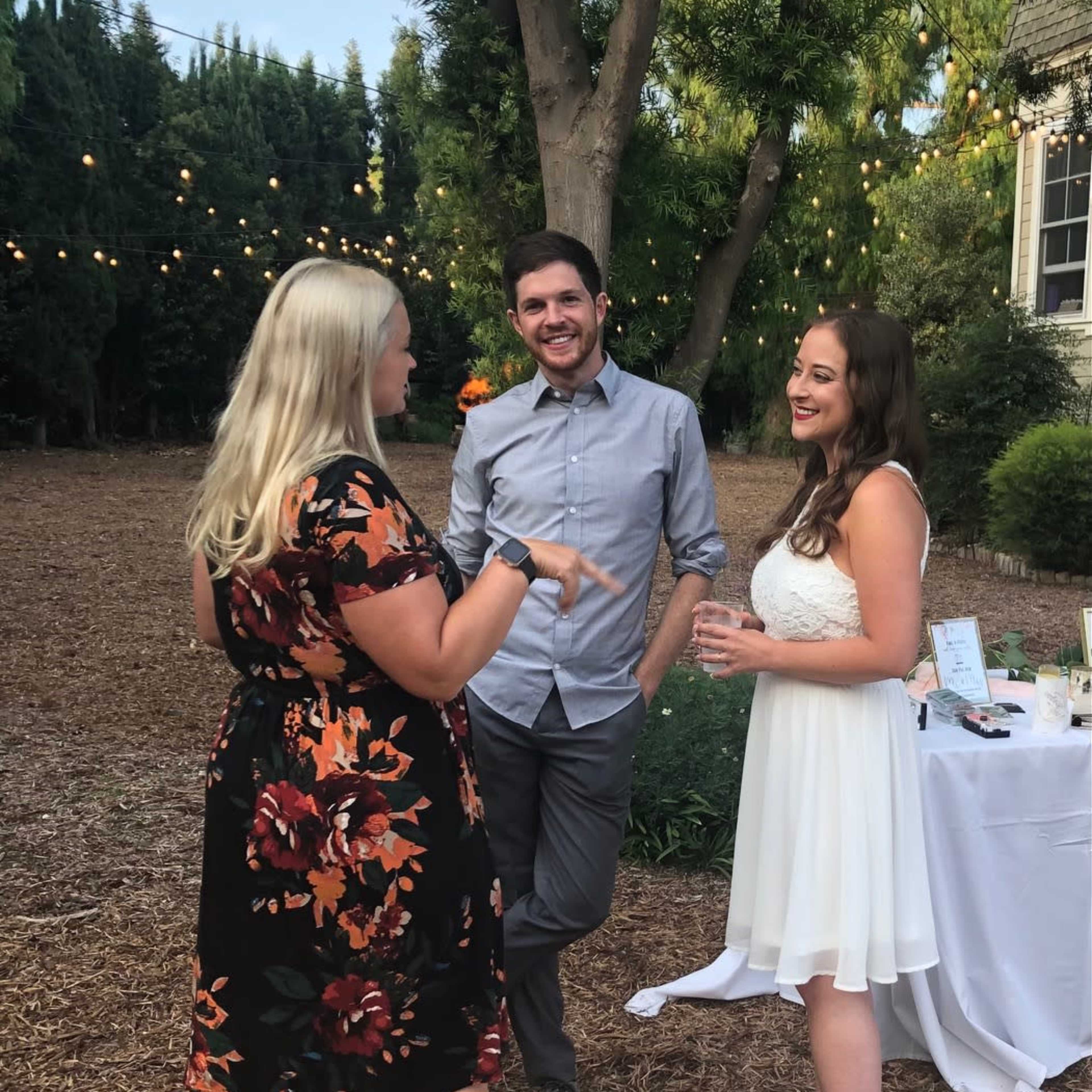 Three people are engaging in conversation outdoors at a gathering, with string lights visible in the background.