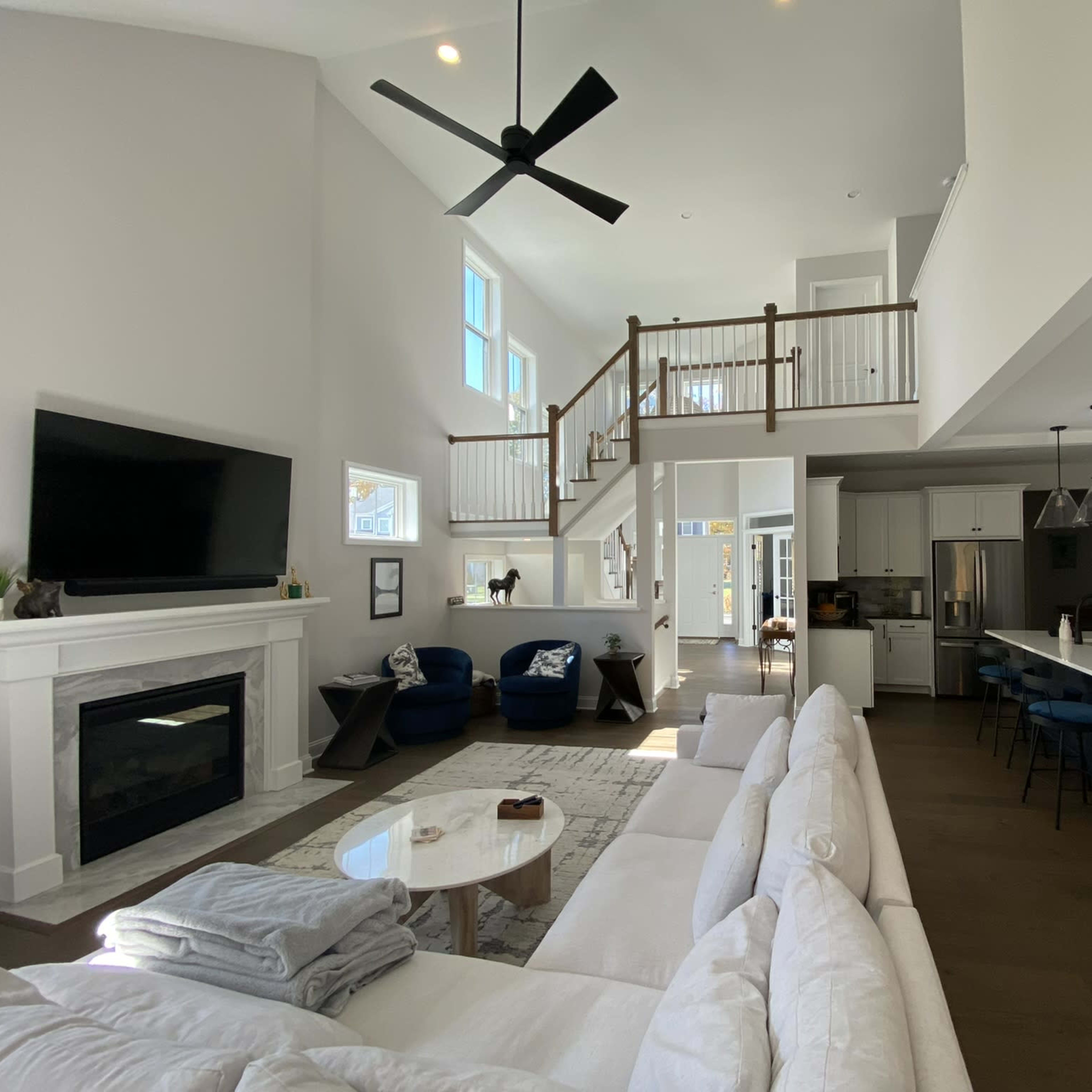 A spacious and modern living area with a white sectional sofa, a large flat-screen TV mounted above a fireplace, and an open kitchen featuring an island with bar seating.