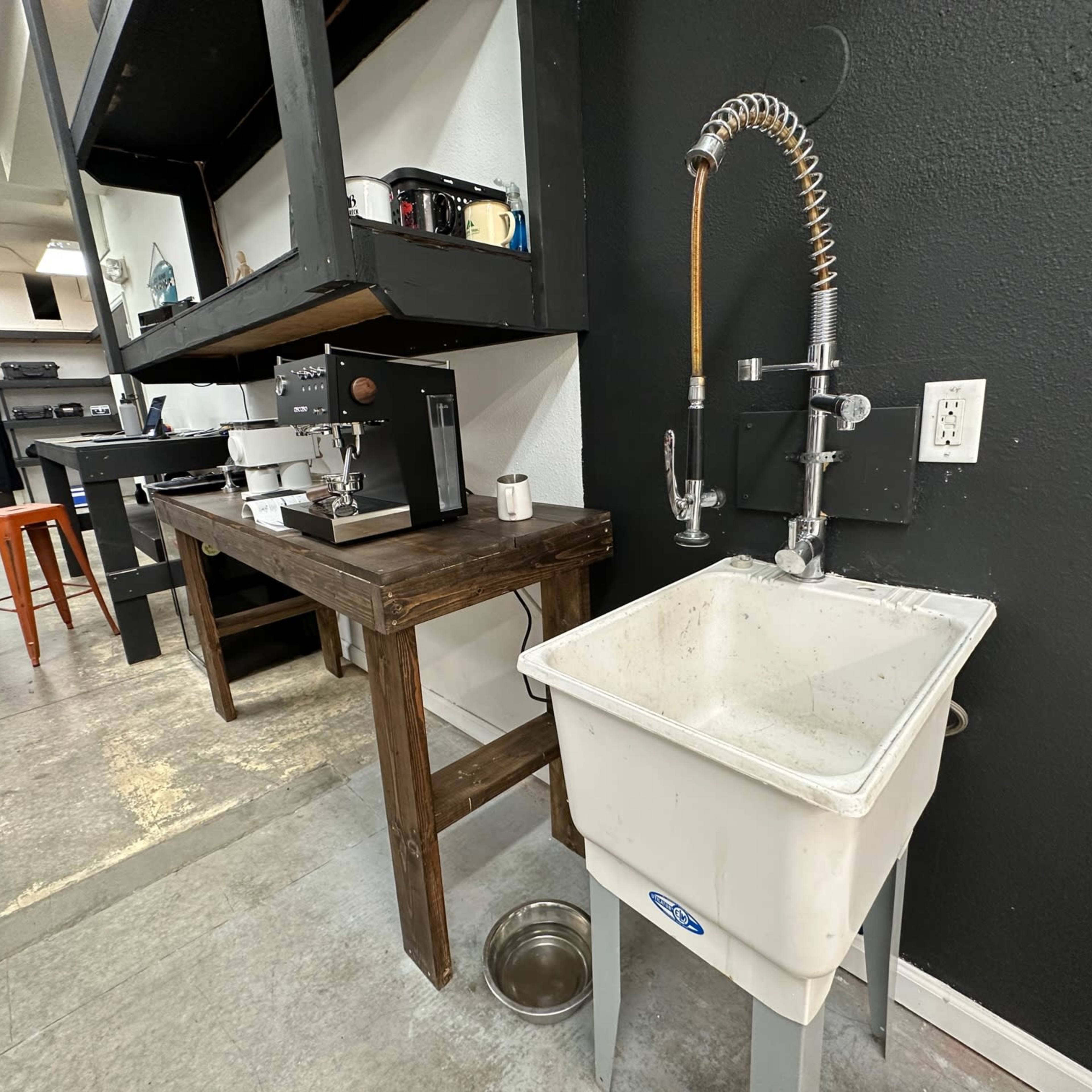 A white utility sink next to a coffee machine and wooden table in a minimalist workspace.