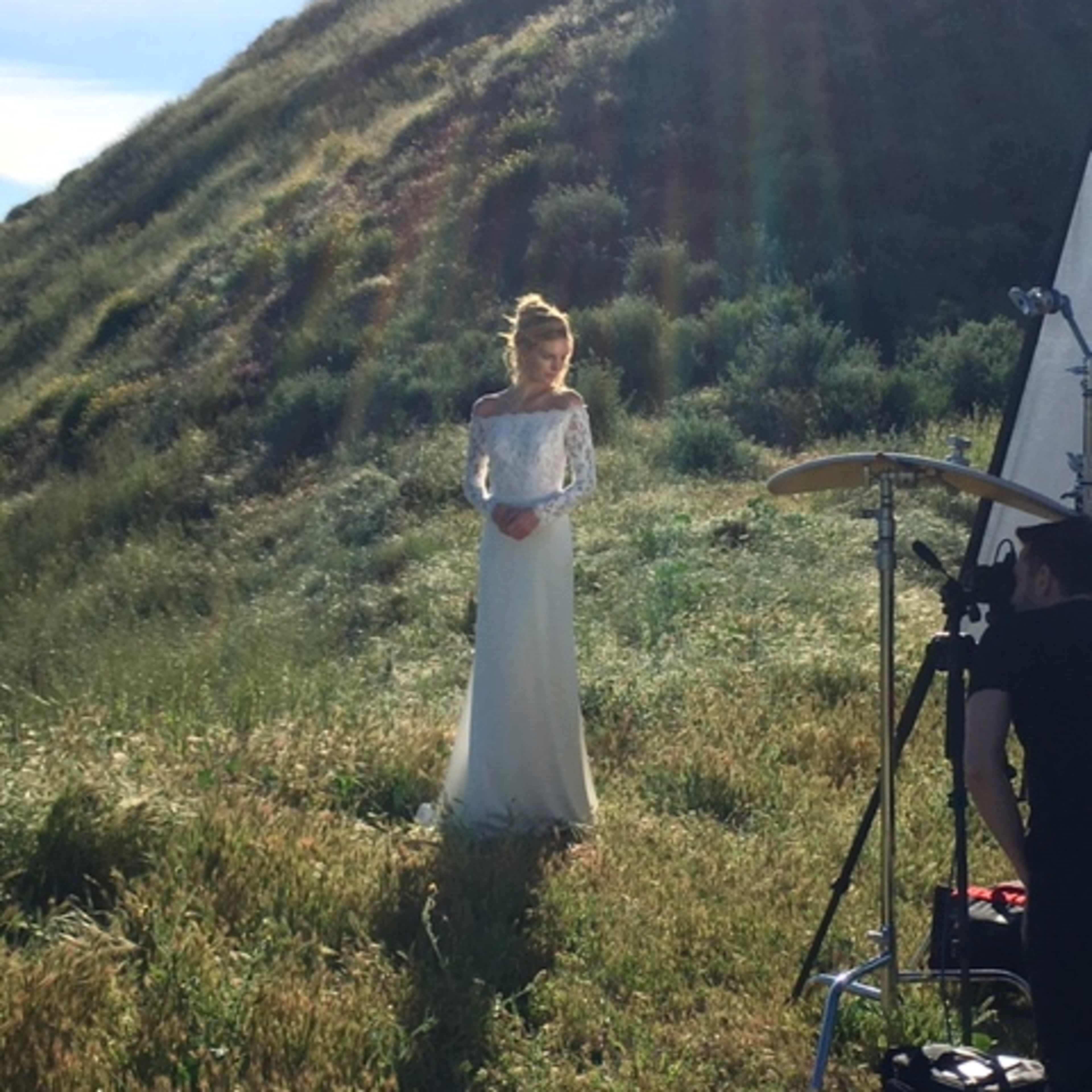 A woman in a long white dress stands on a grassy hillside while a photographer sets up equipment nearby.