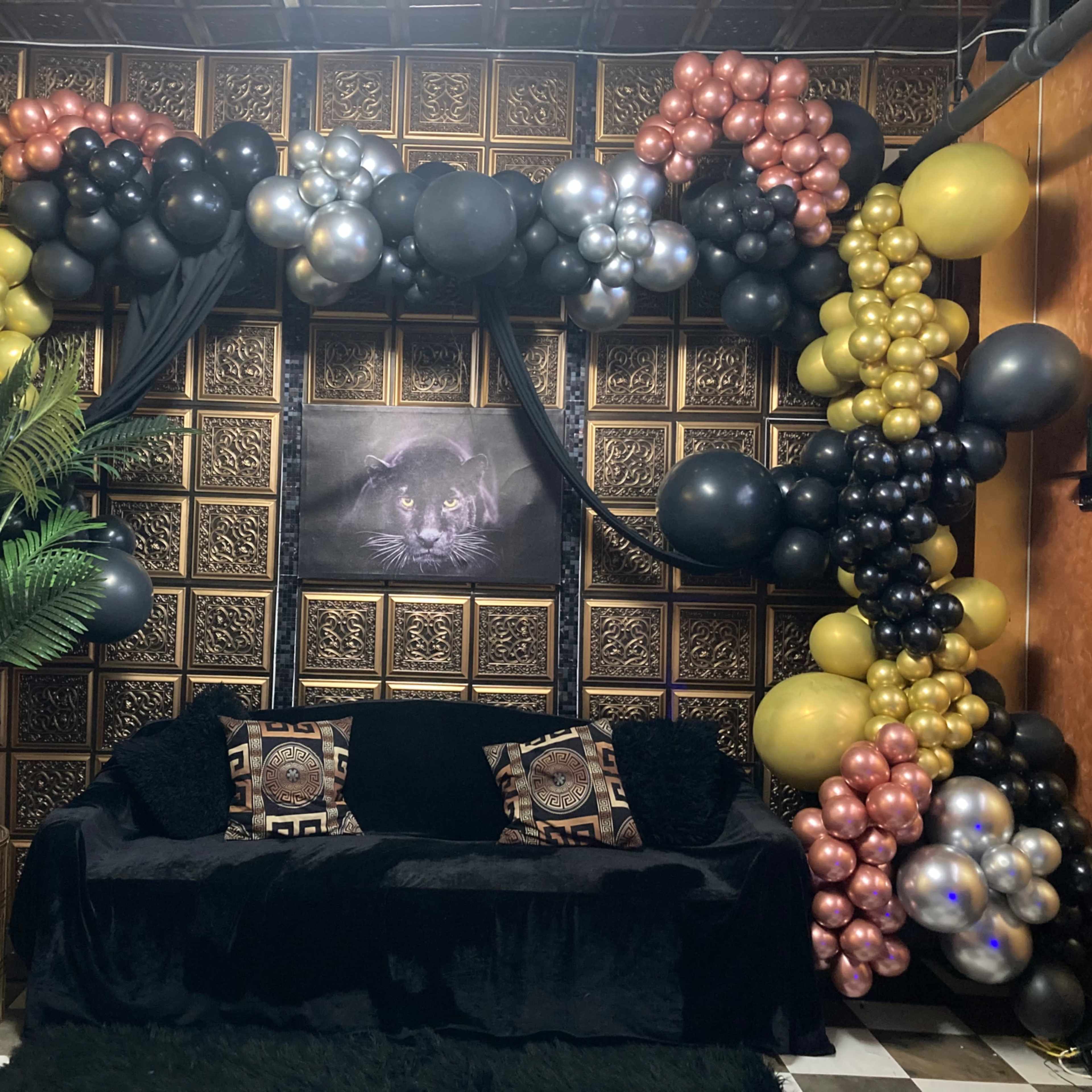 A decorated corner of a room featuring a black couch with decorative pillows, a large plant, and an archway of balloons in black, gold, and silver against a textured wall.