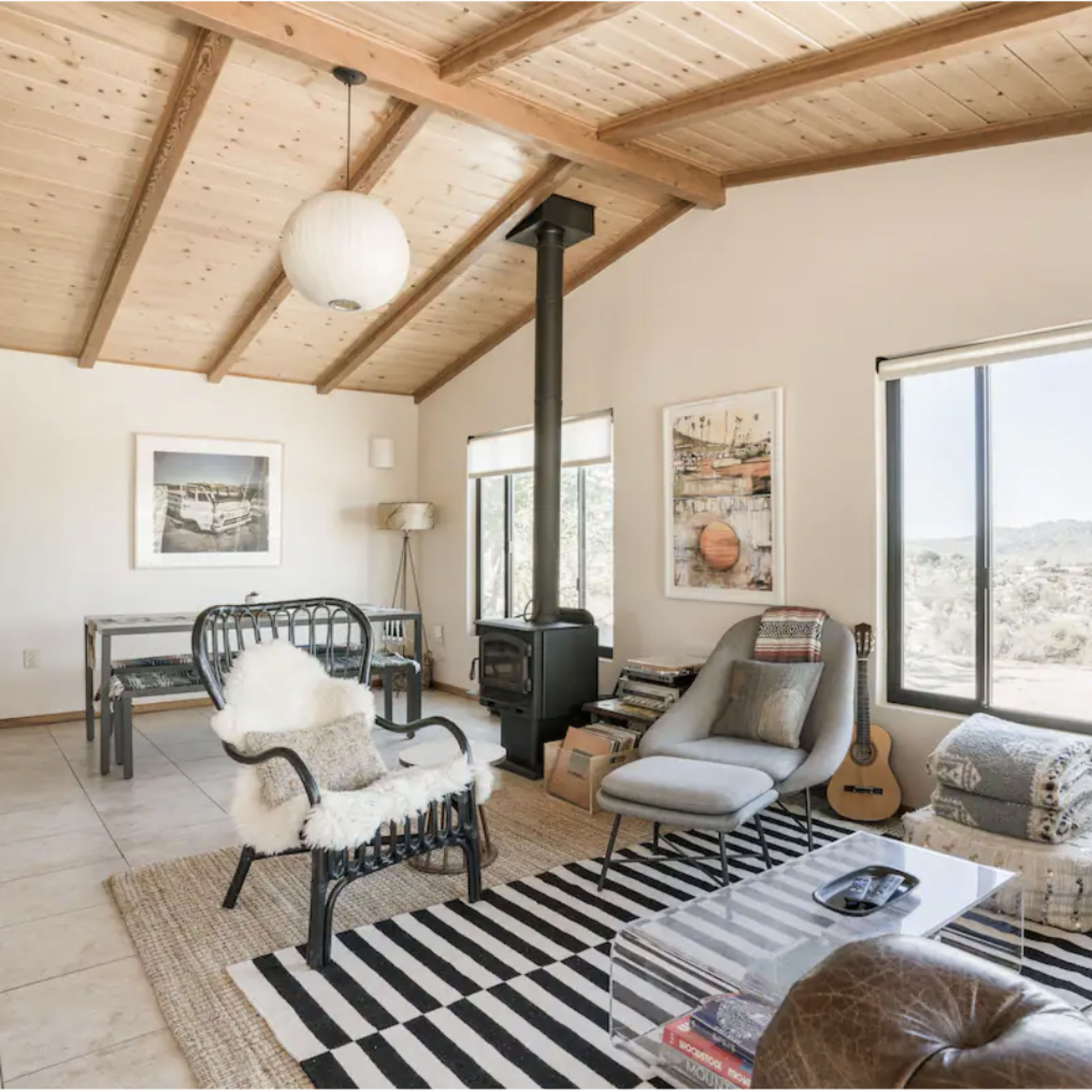 A spacious living area with a wooden ceiling, large windows, and a mix of modern and rustic furniture, including a glass coffee table and a guitar.