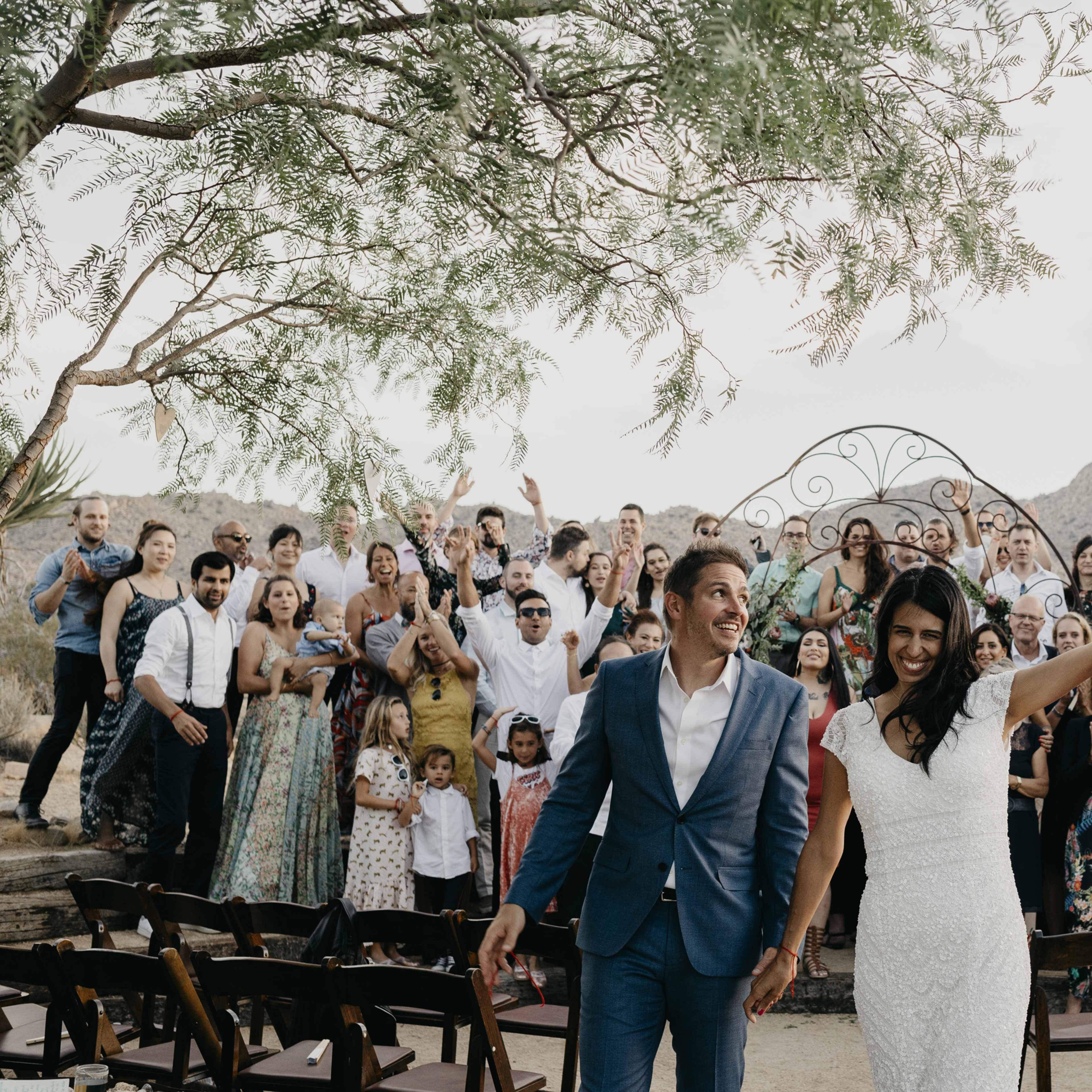 A newlywed couple walks hand in hand, smiling and celebrating, as their guests stand behind them, cheering in a desert landscape.