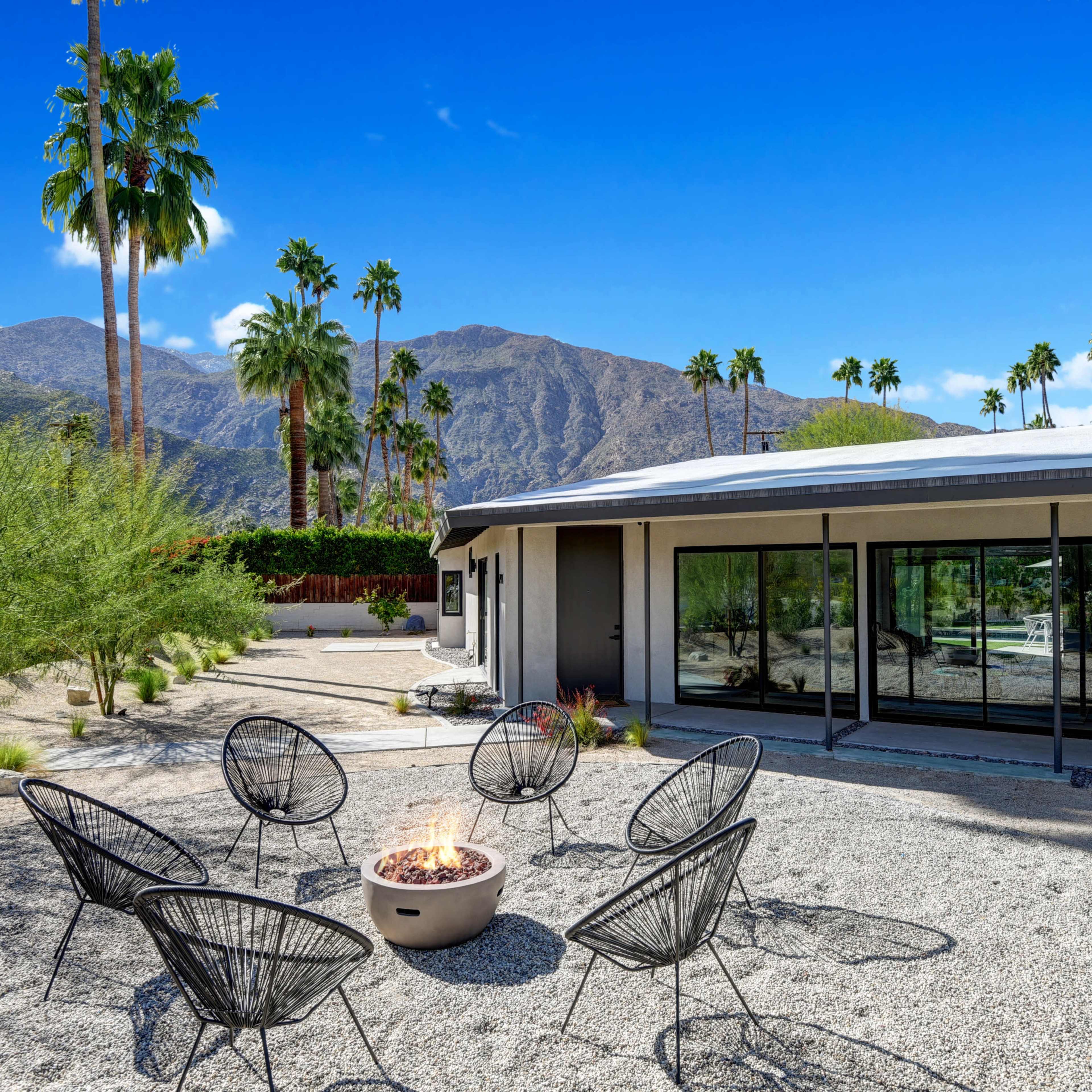 A modern outdoor space features six black chairs arranged around a fire pit, with palm trees and mountains in the background.