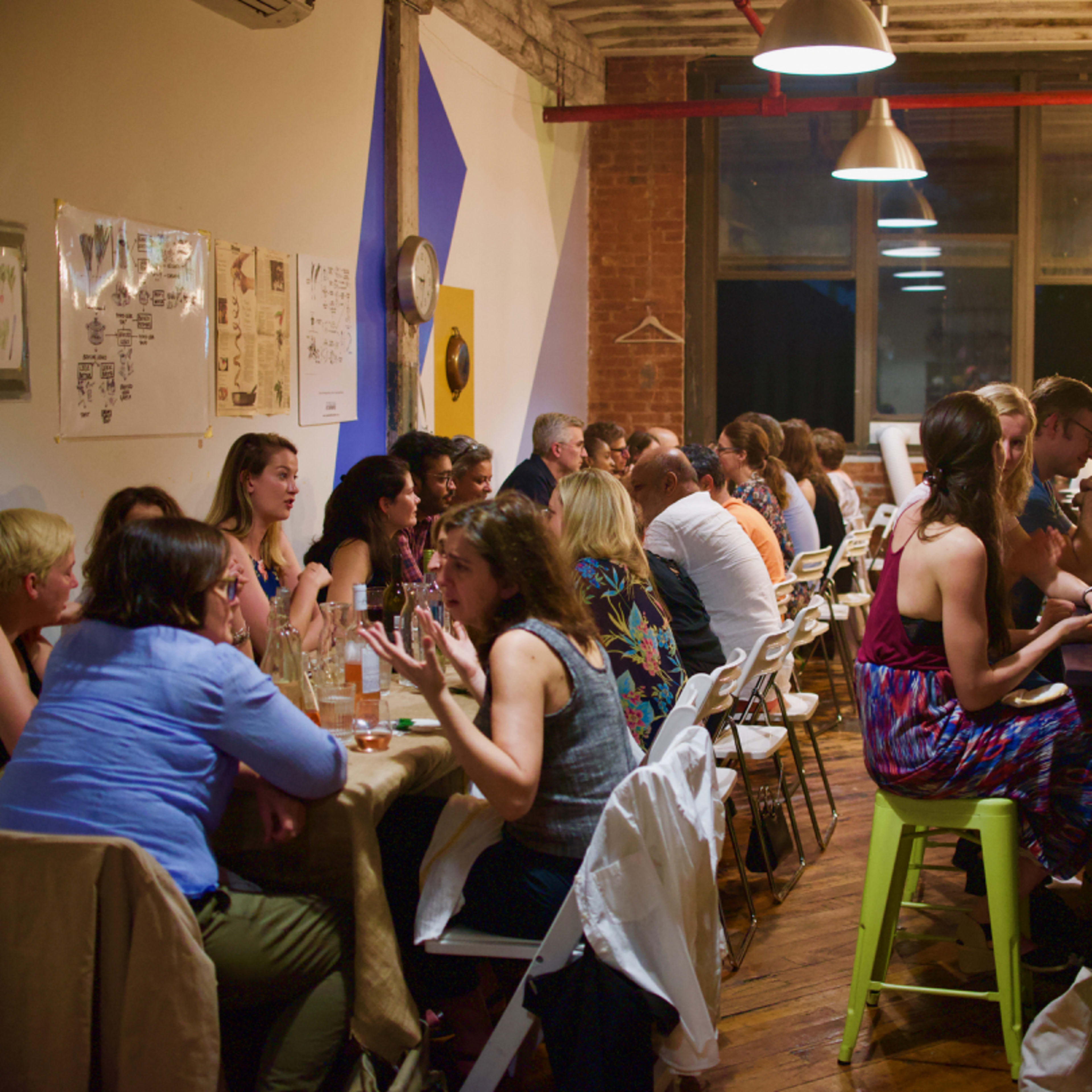 A group of people is seated at long tables in a dimly lit restaurant, engaged in conversation and enjoying their meals.