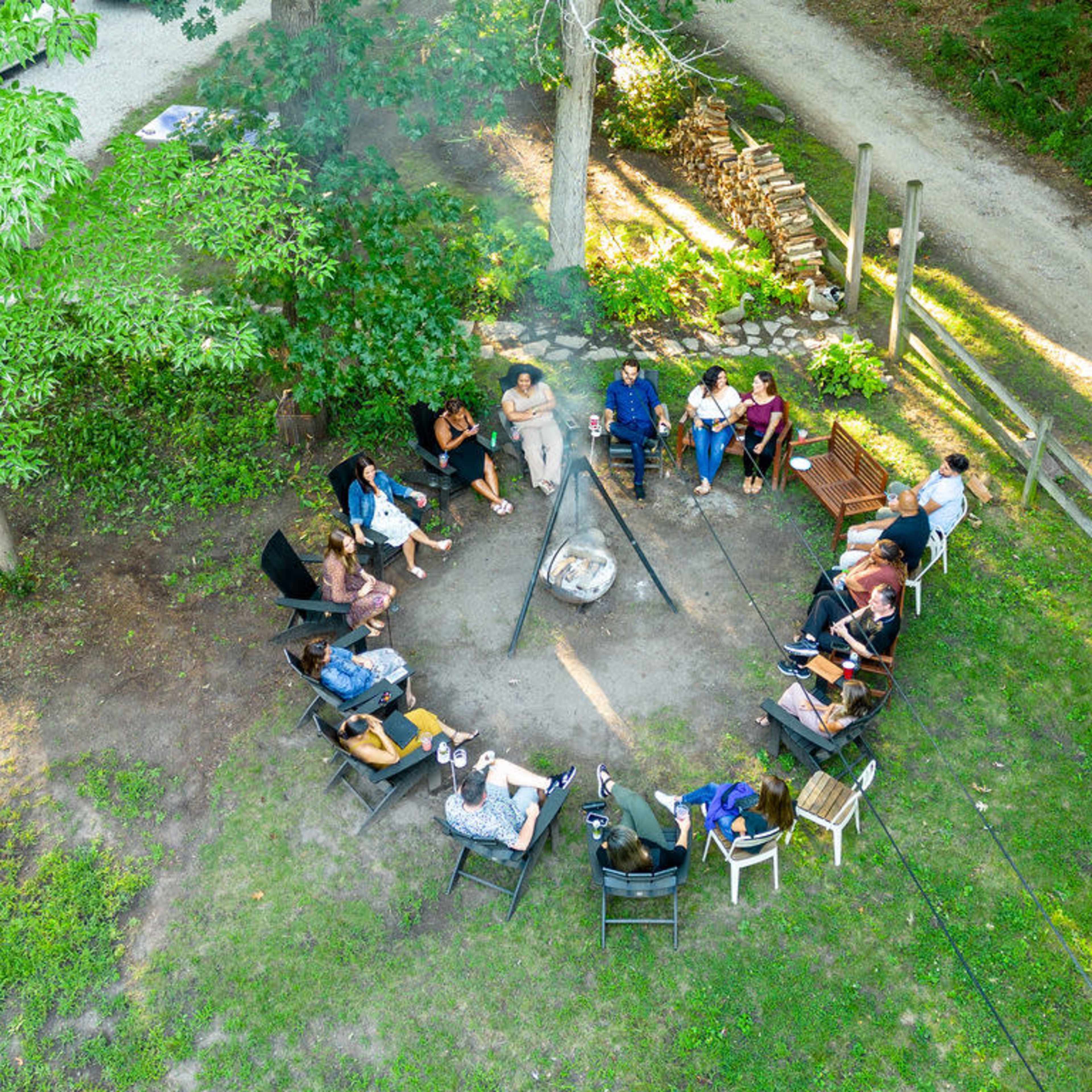 A group of people sit in a circle around a campfire in a grassy area surrounded by trees.