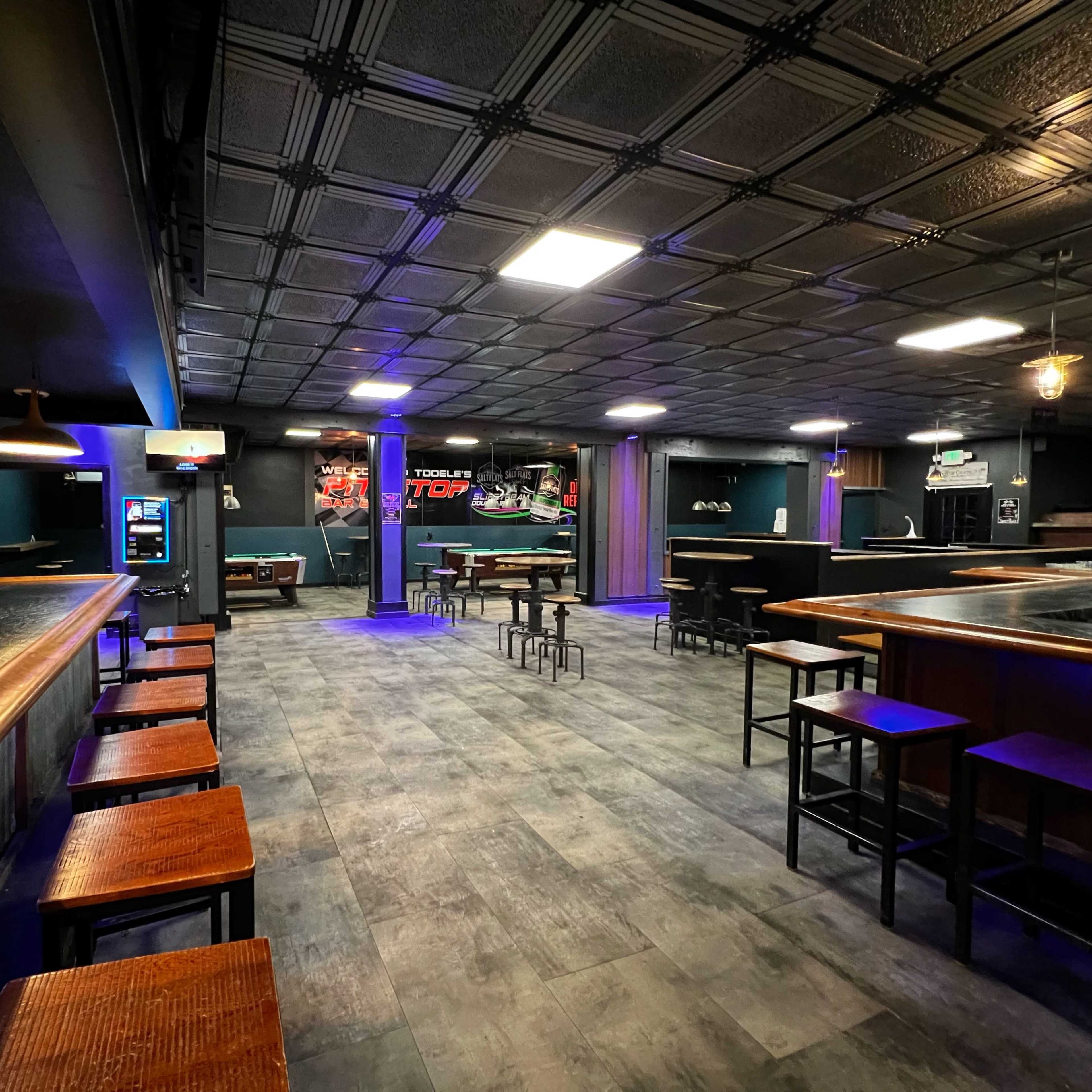 An empty bar area with a long counter, high tables, and dark walls, illuminated by blue and white lights.