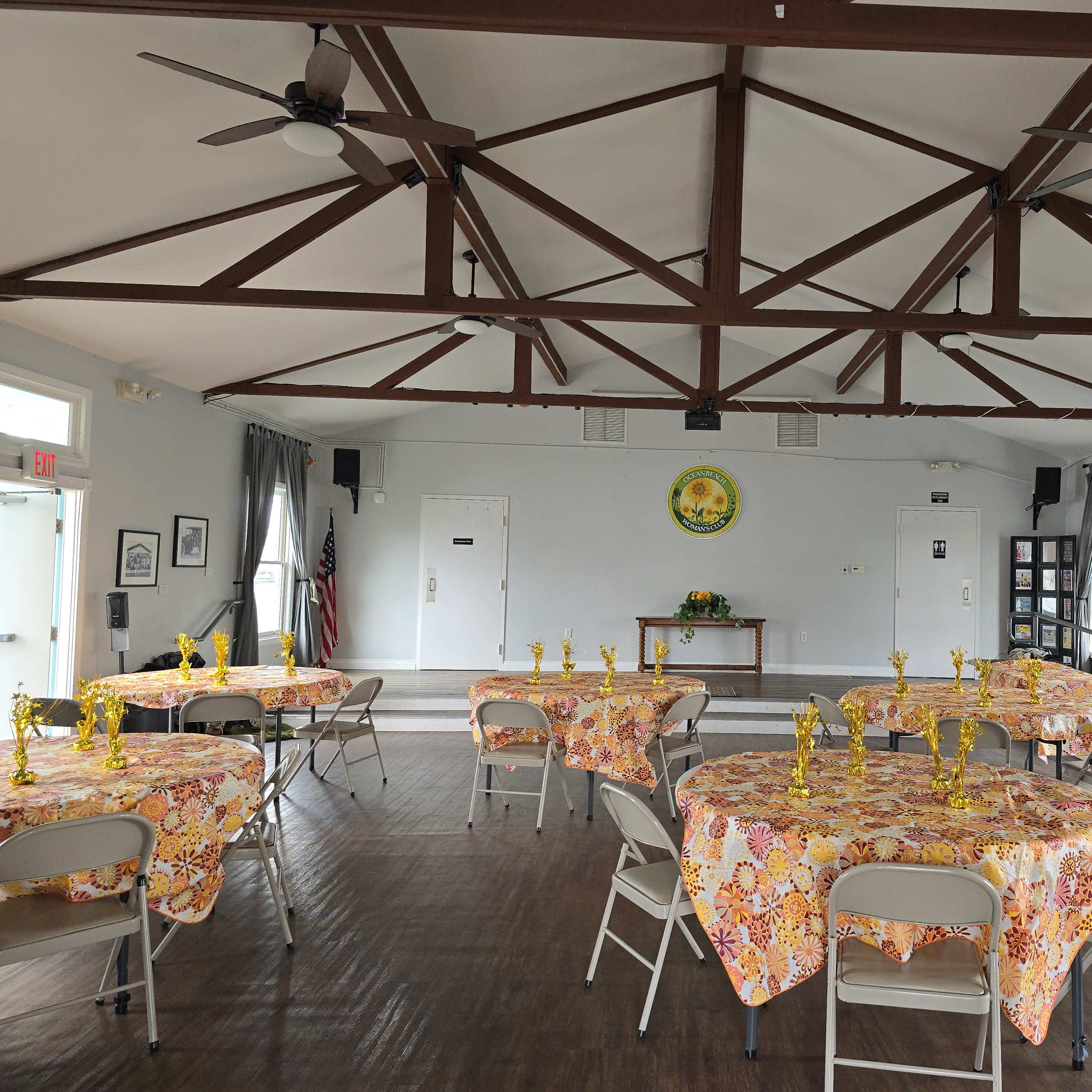 A banquet hall with several round tables covered in floral tablecloths, each adorned with golden candle holders, and a podium at the front of the room.