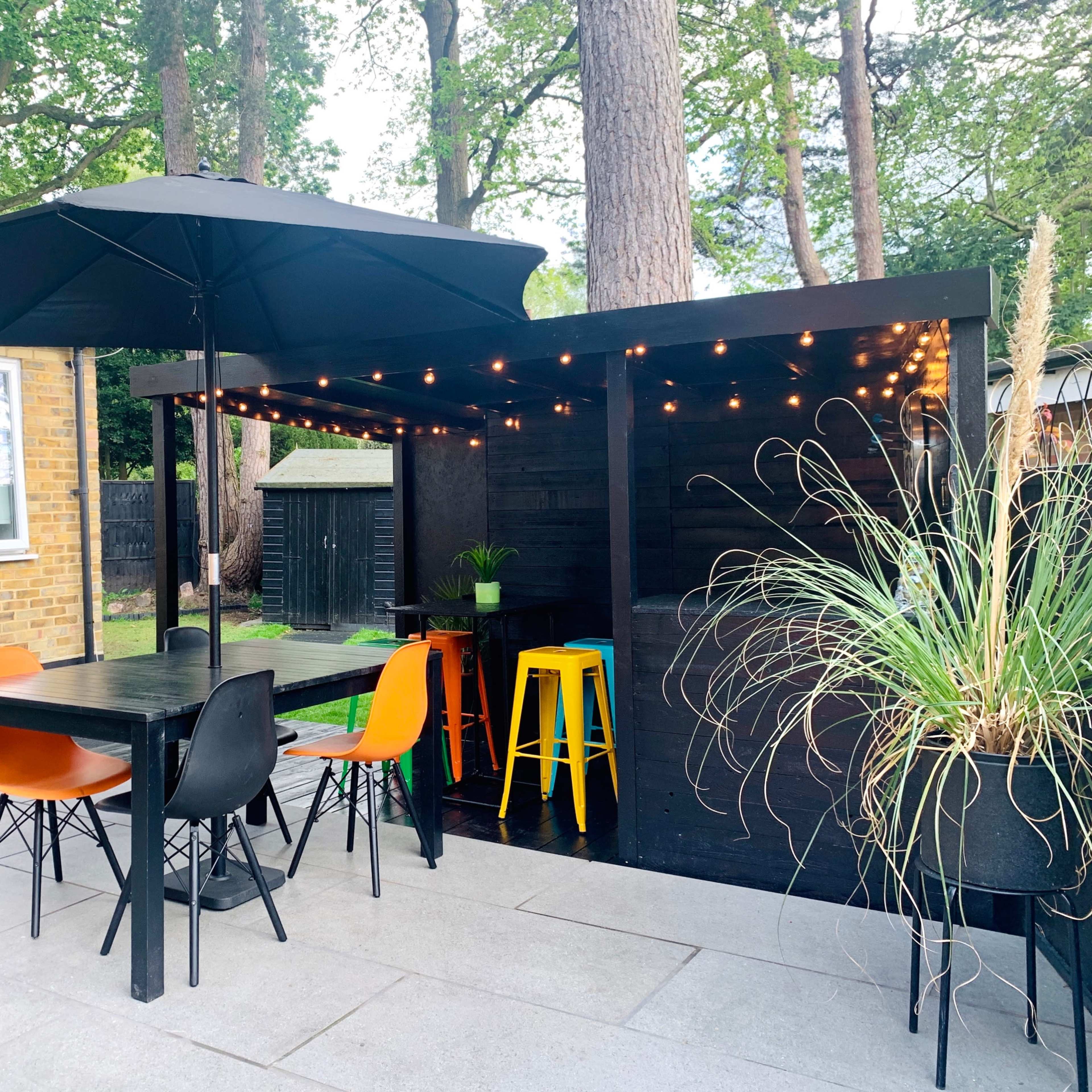 A backyard patio features a black wooden structure with string lights, an umbrella, and colorful chairs around a black table.