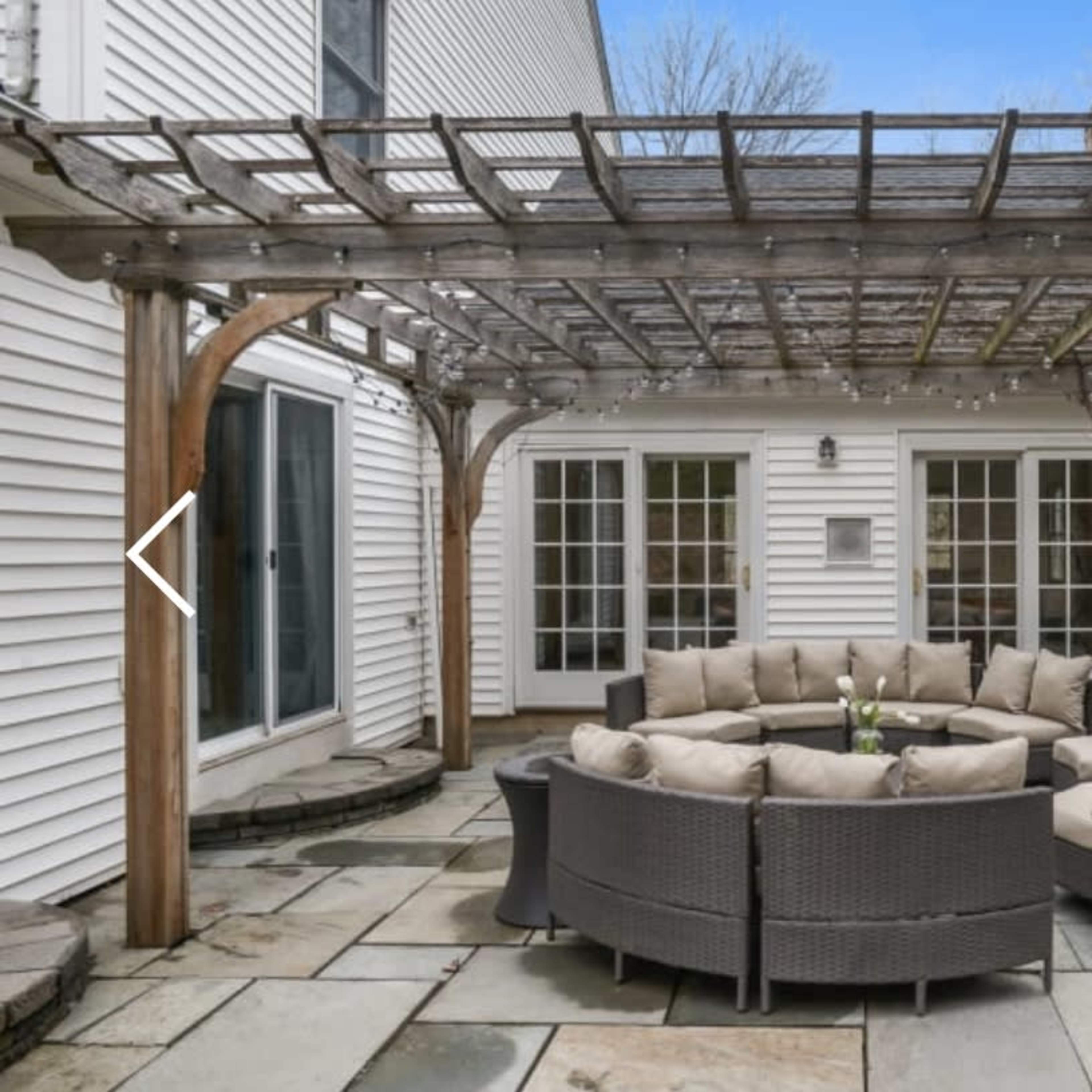 A circular seating area with cushioned chairs is placed under a wooden pergola on a stone patio.