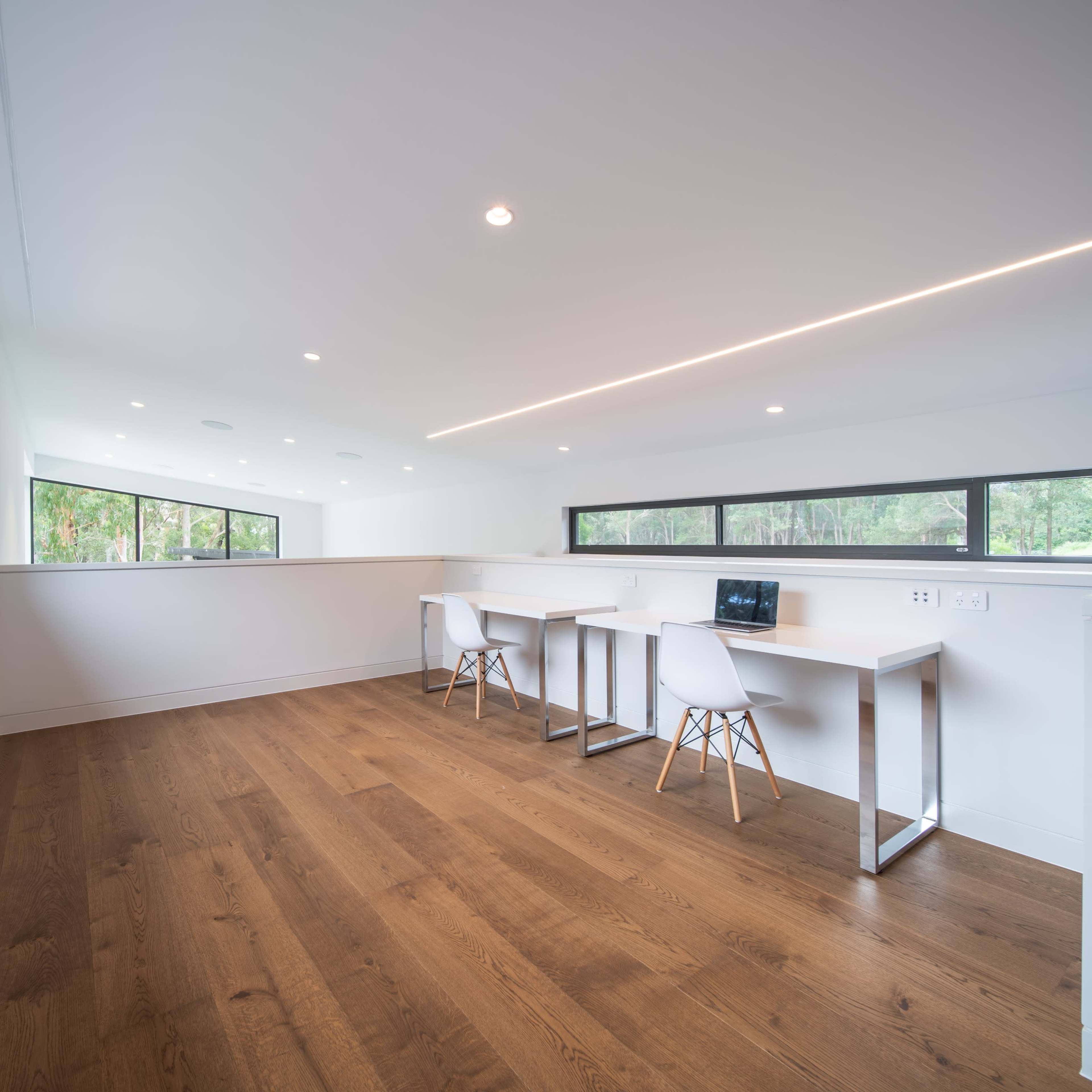 A light-filled, modern workspace featuring two white desks with chairs against a backdrop of large windows.