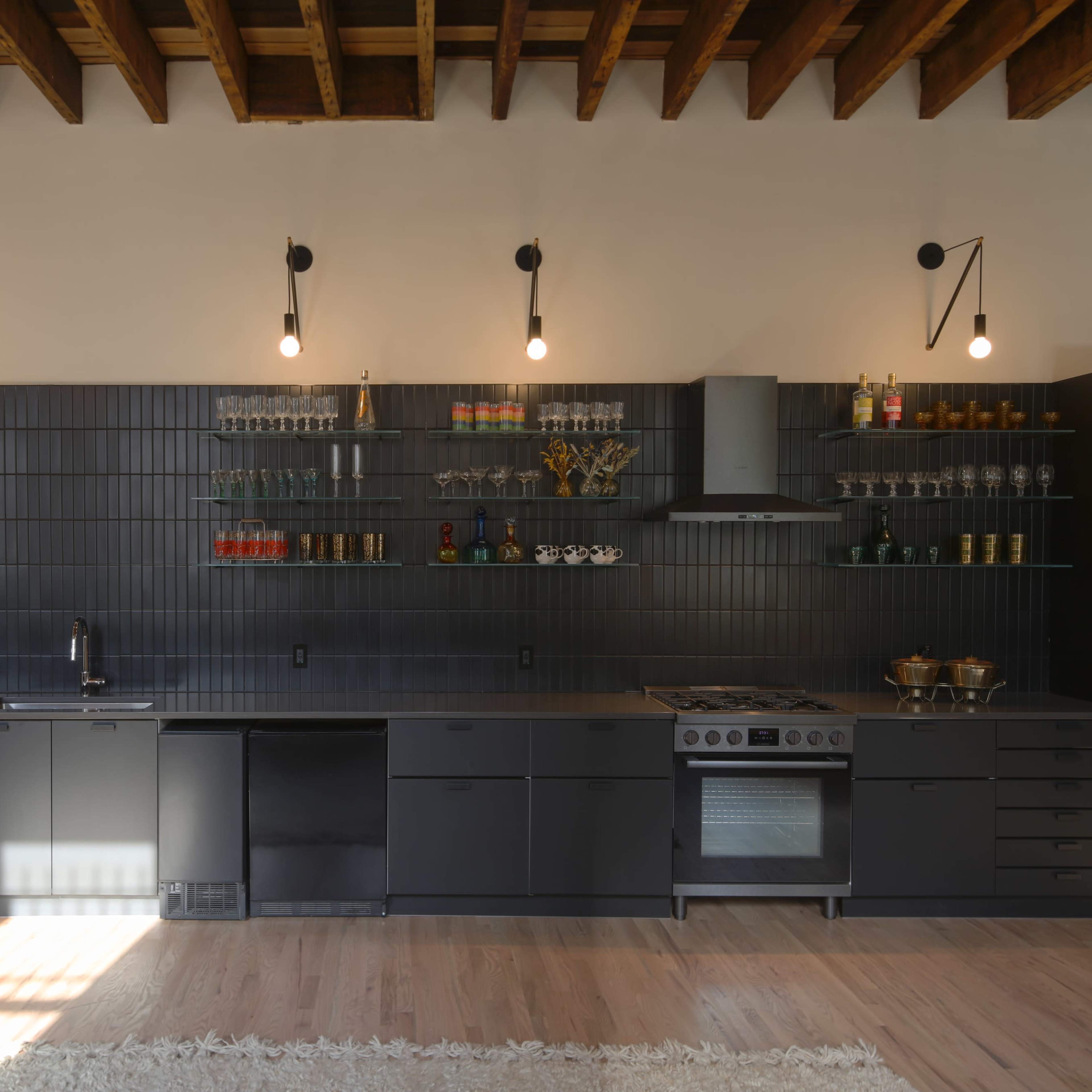 A modern kitchen with a dark themed design, including a long countertop, wall-mounted shelves holding glassware, and professional appliances.