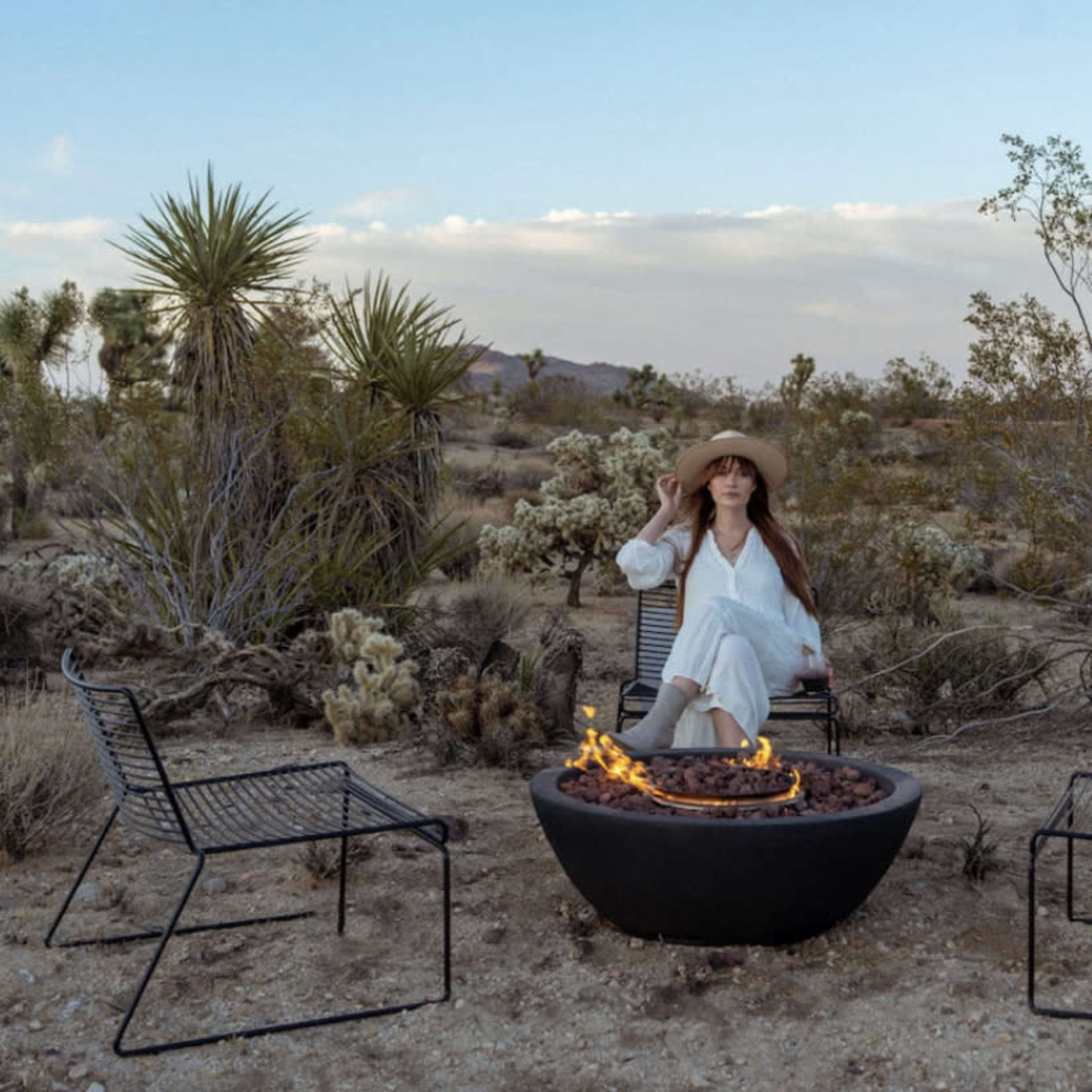 A woman in a wide-brimmed hat sits in a chair near a fire pit surrounded by desert vegetation and sparse bushes.