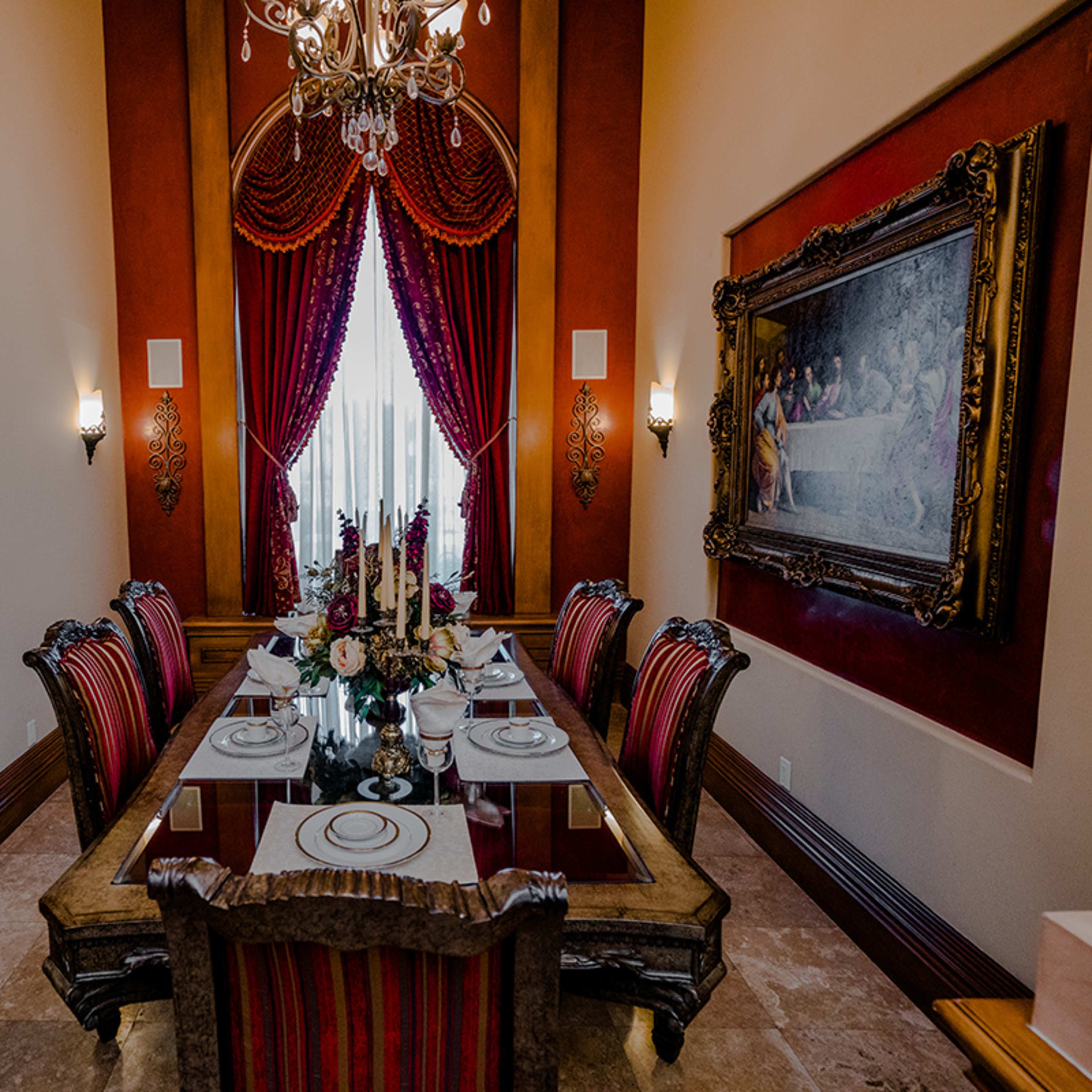 A formal dining room features a long table set for a meal, surrounded by red-striped chairs, with elegant drapes and a chandelier above.