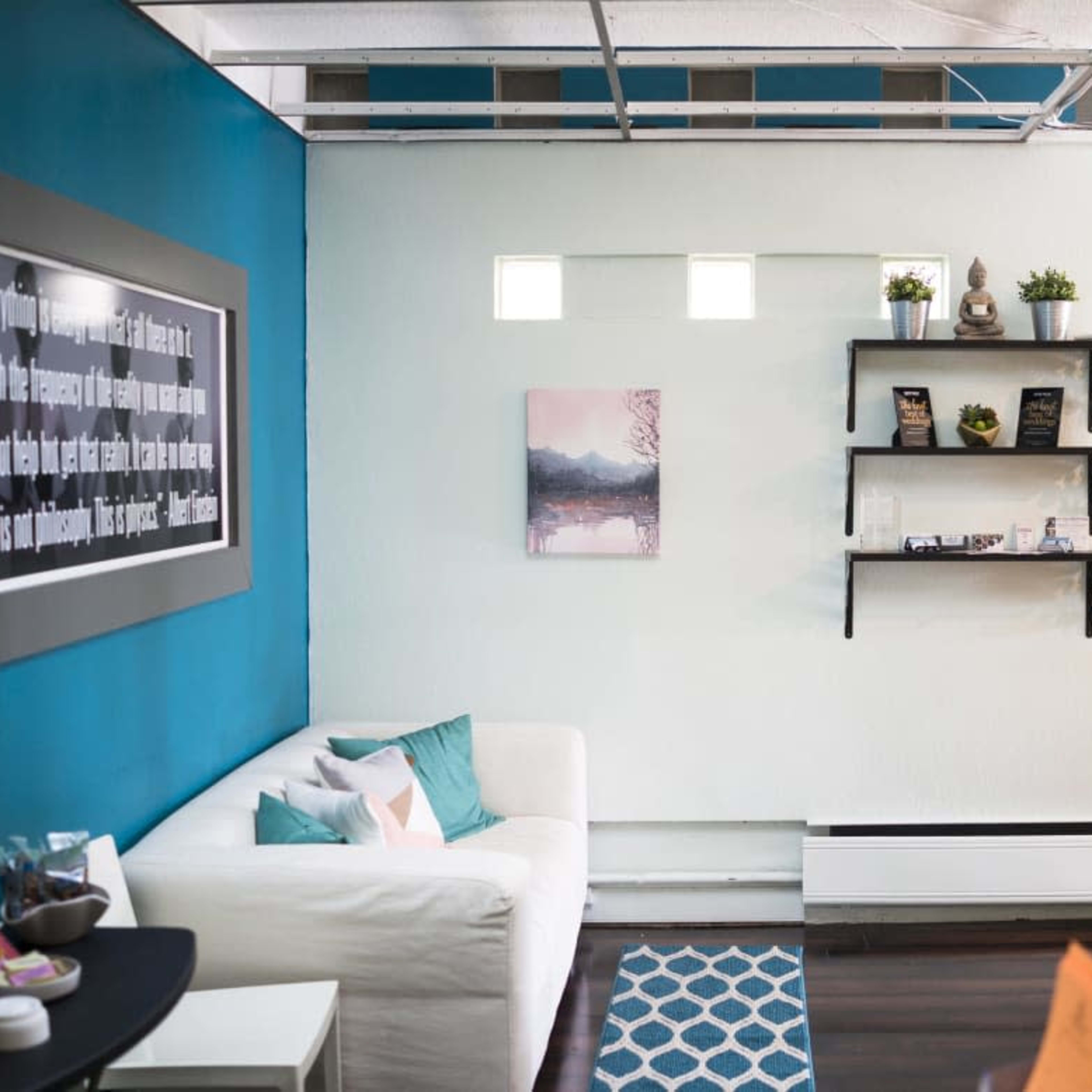 A cozy room with a white sofa, teal walls, a small shelf displaying plants and decorative items, and a piece of art on the wall.