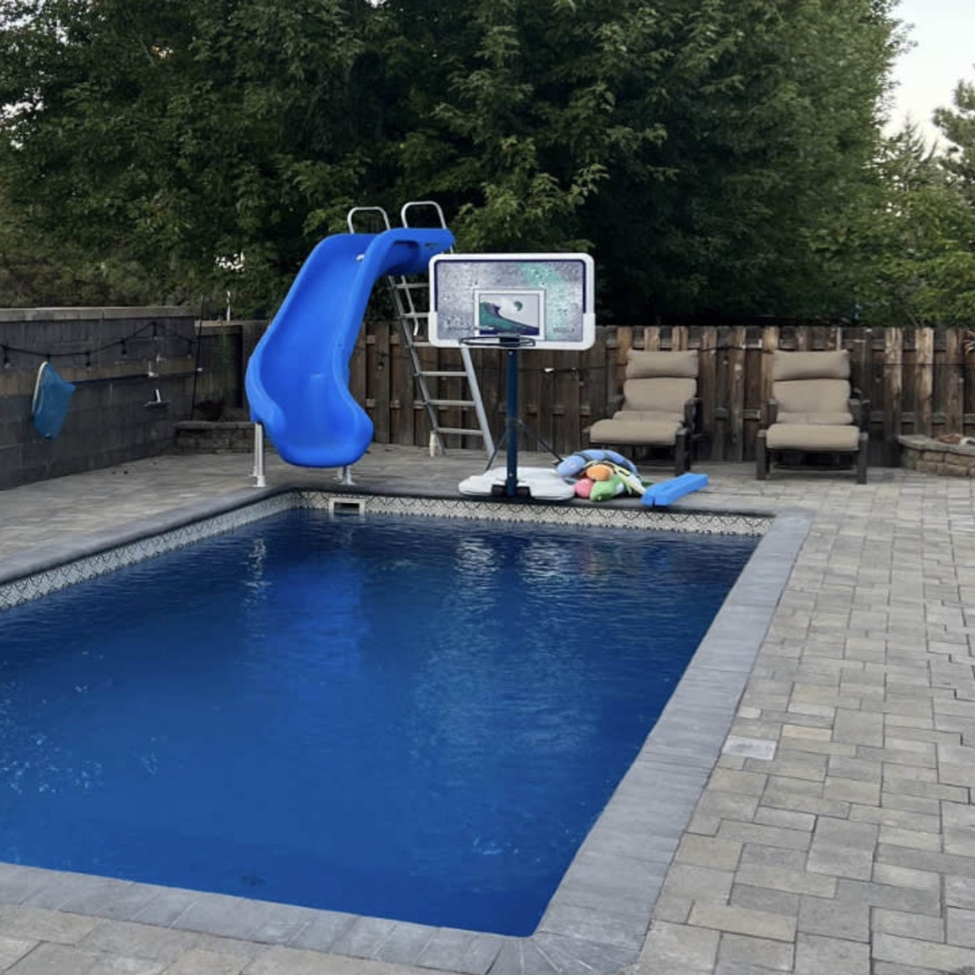 A backyard pool with a blue waterslide, a basketball hoop, and lounge chairs arranged beside the water.