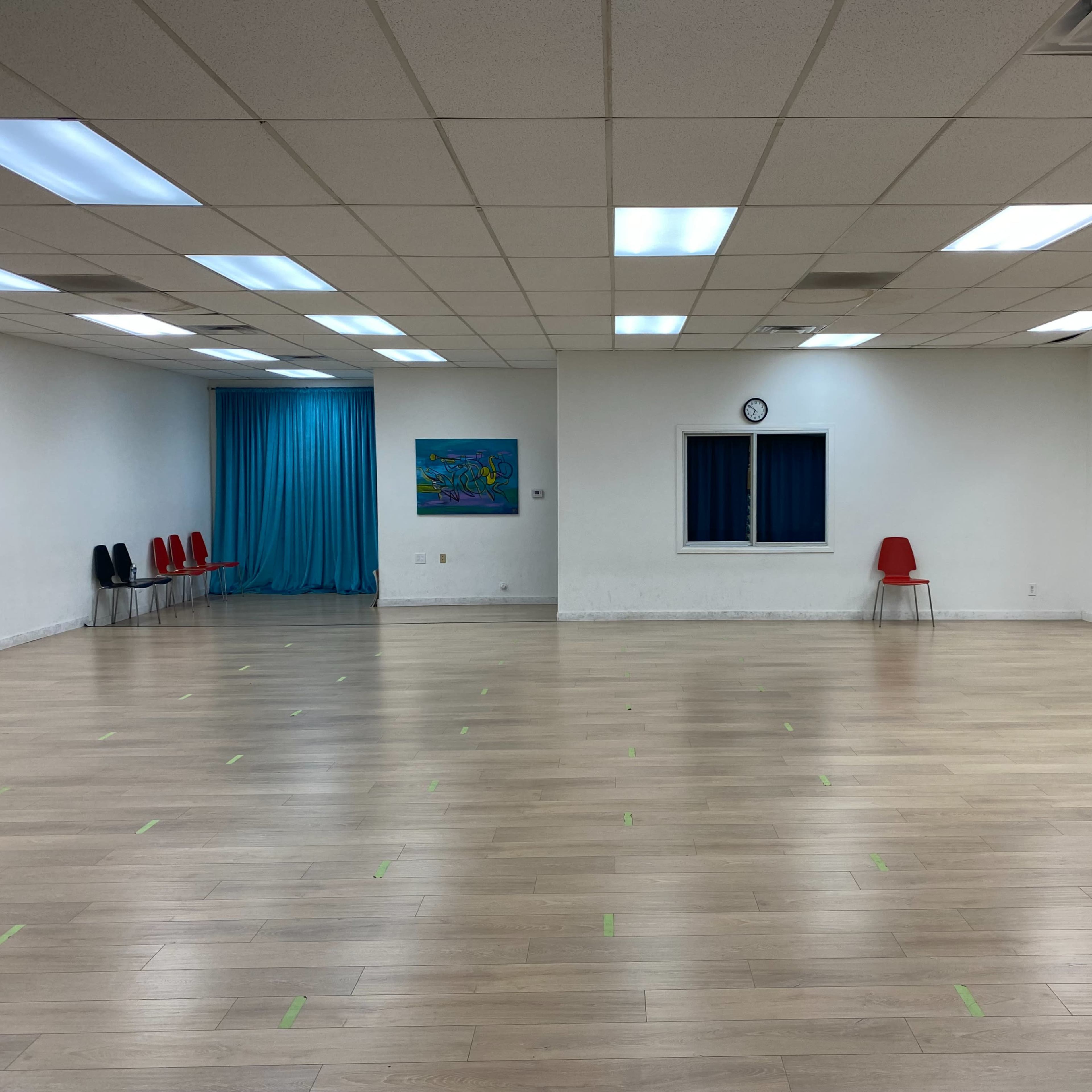 A spacious dance studio featuring a wooden floor, a large mirror, red chairs along the walls, and a blue curtain in the background.