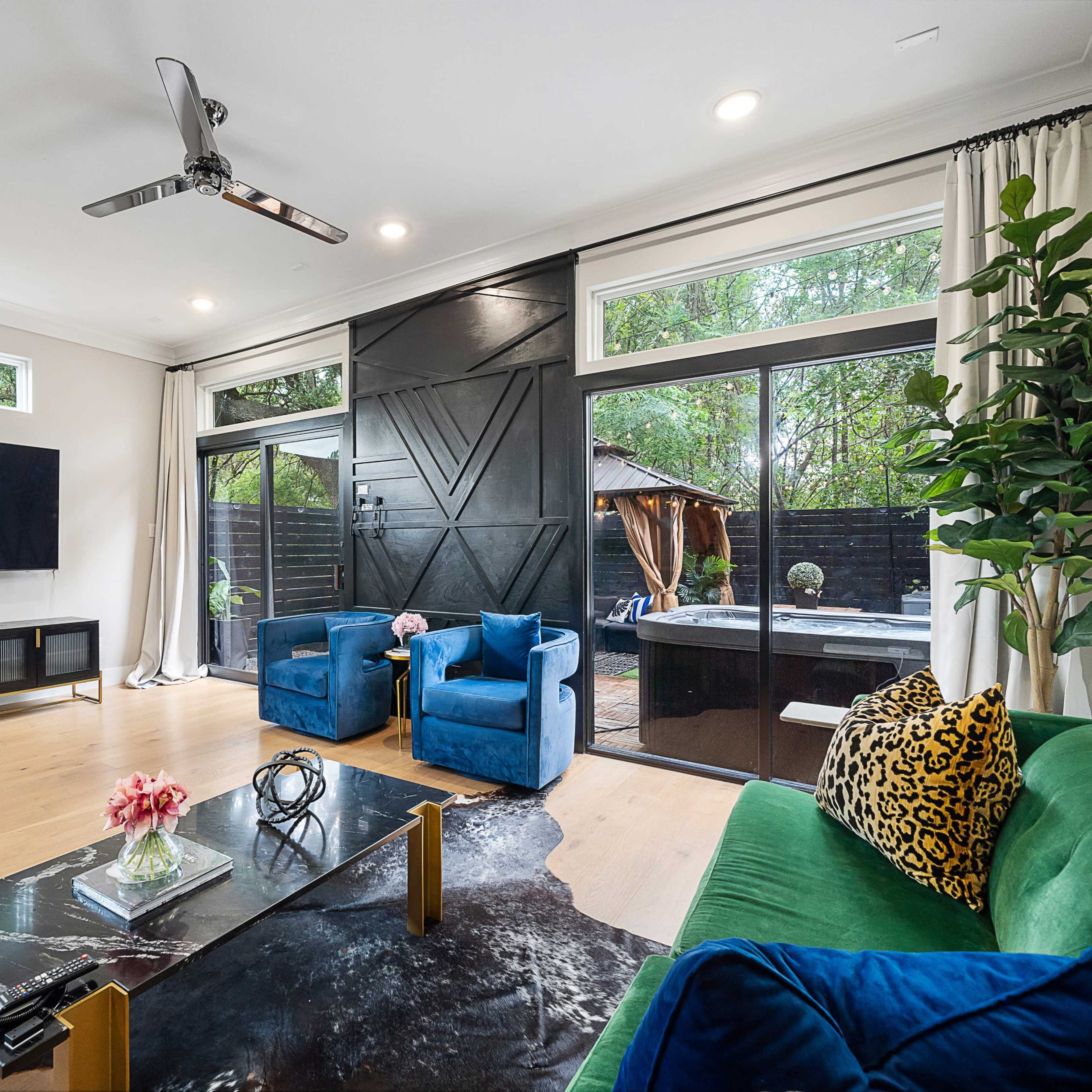 A modern living room with large windows, featuring a green sofa, blue chairs, a coffee table, and a wall-mounted television, surrounded by indoor plants.