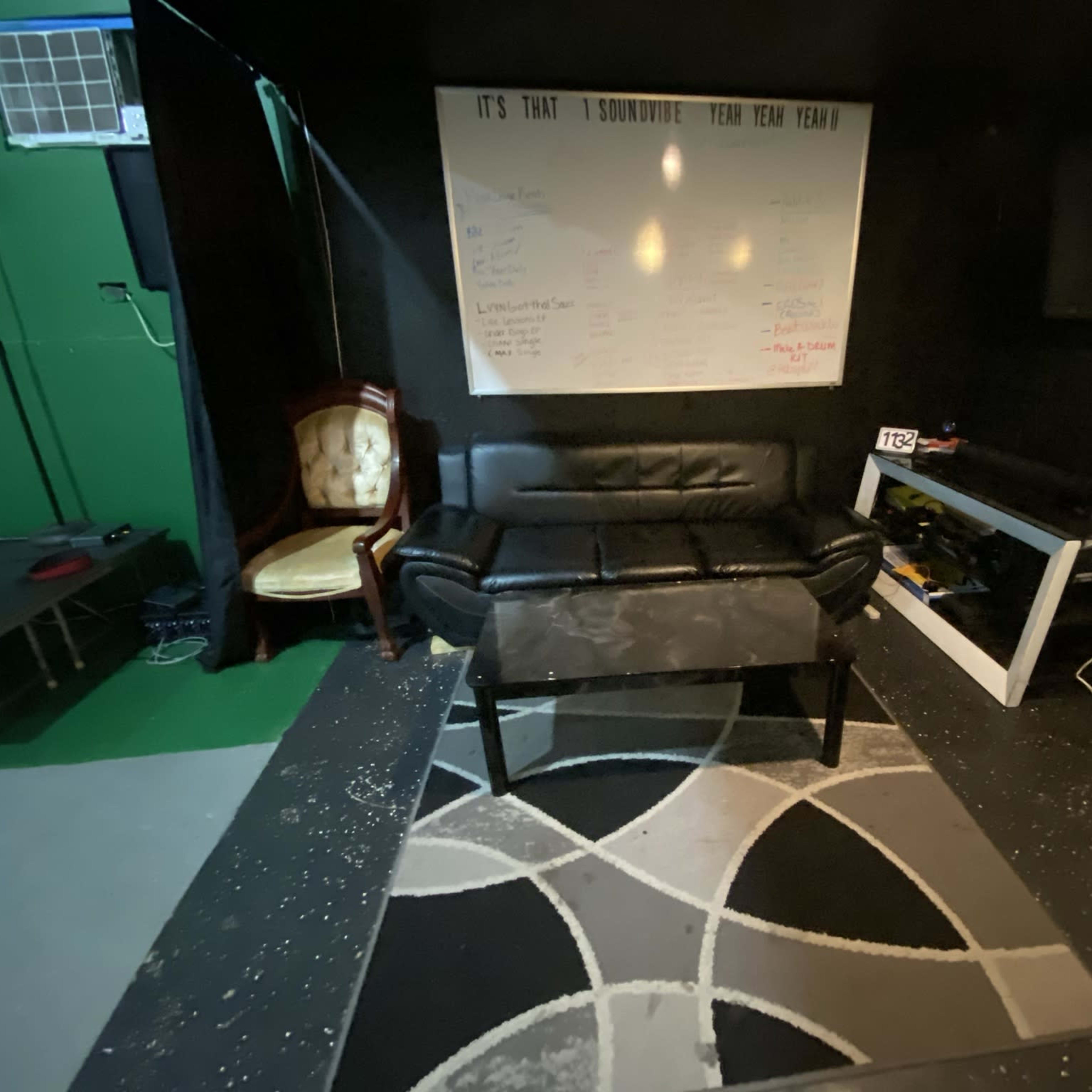 A black leather couch and a coffee table are positioned on a patterned rug in a dimly lit room with a whiteboard and television against one wall.