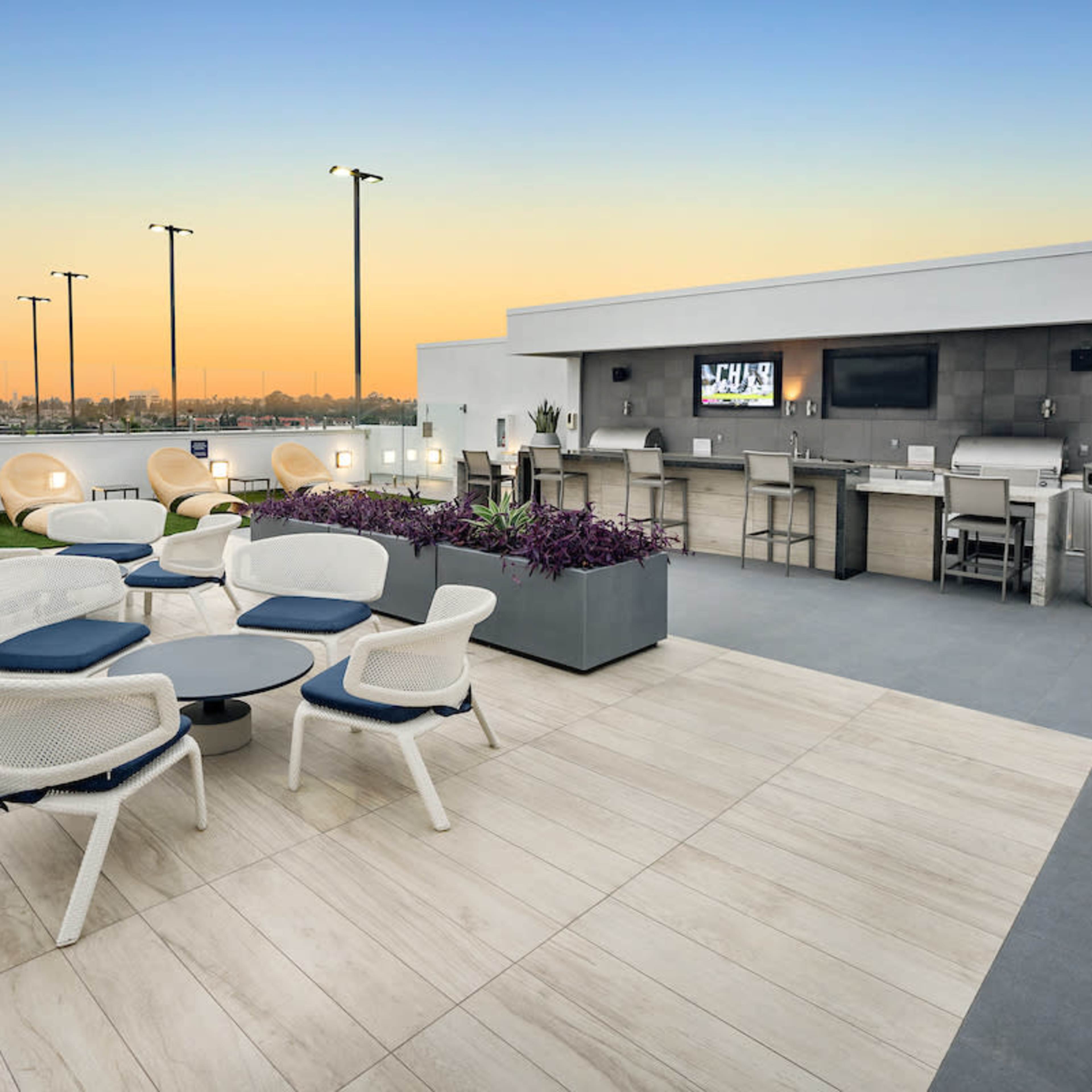 A rooftop terrace featuring modern seating arrangements, a bar area, and a view of the skyline at sunset.