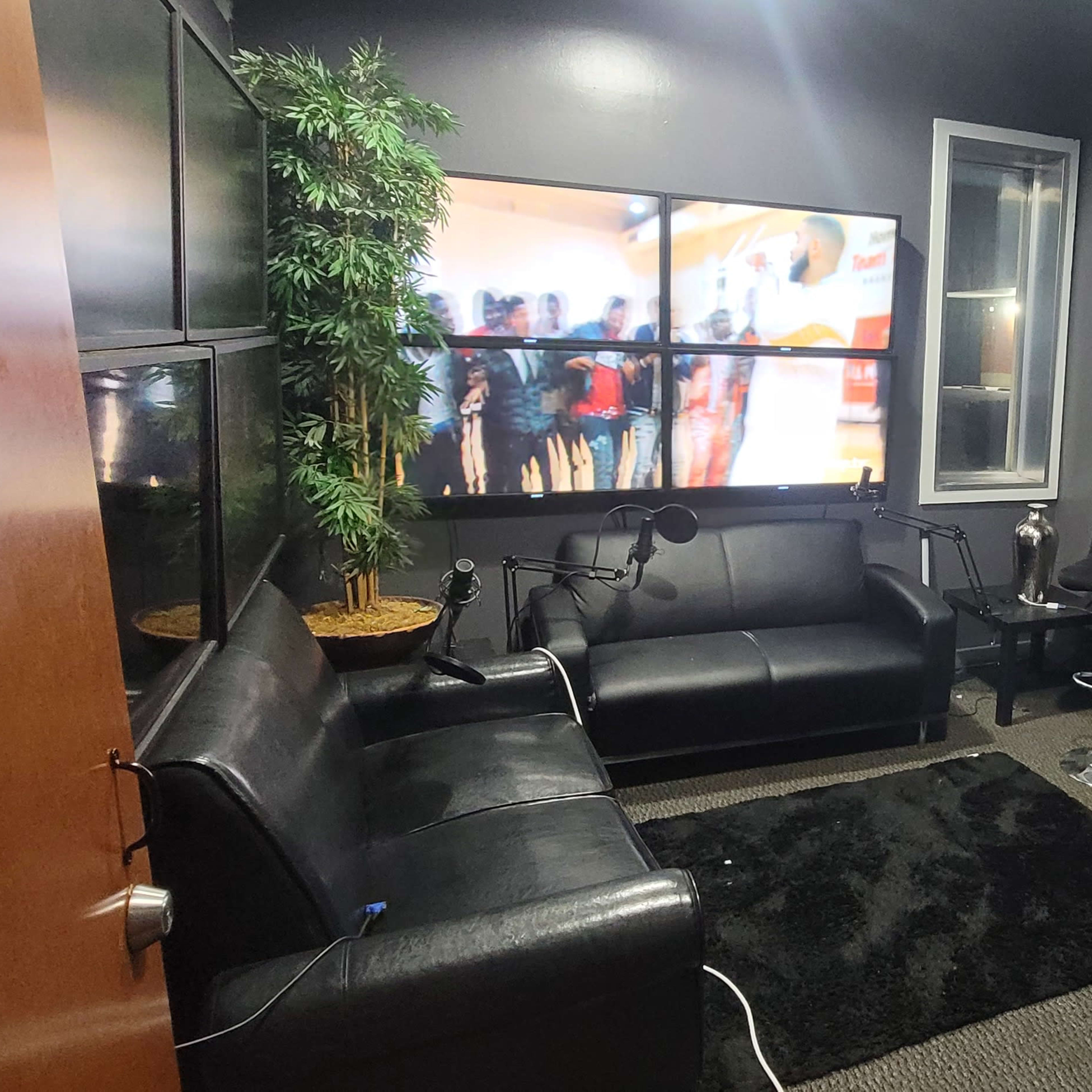A small, modern room furnished with a black leather sofa, bar stools, and a large screen displaying people in another setting.