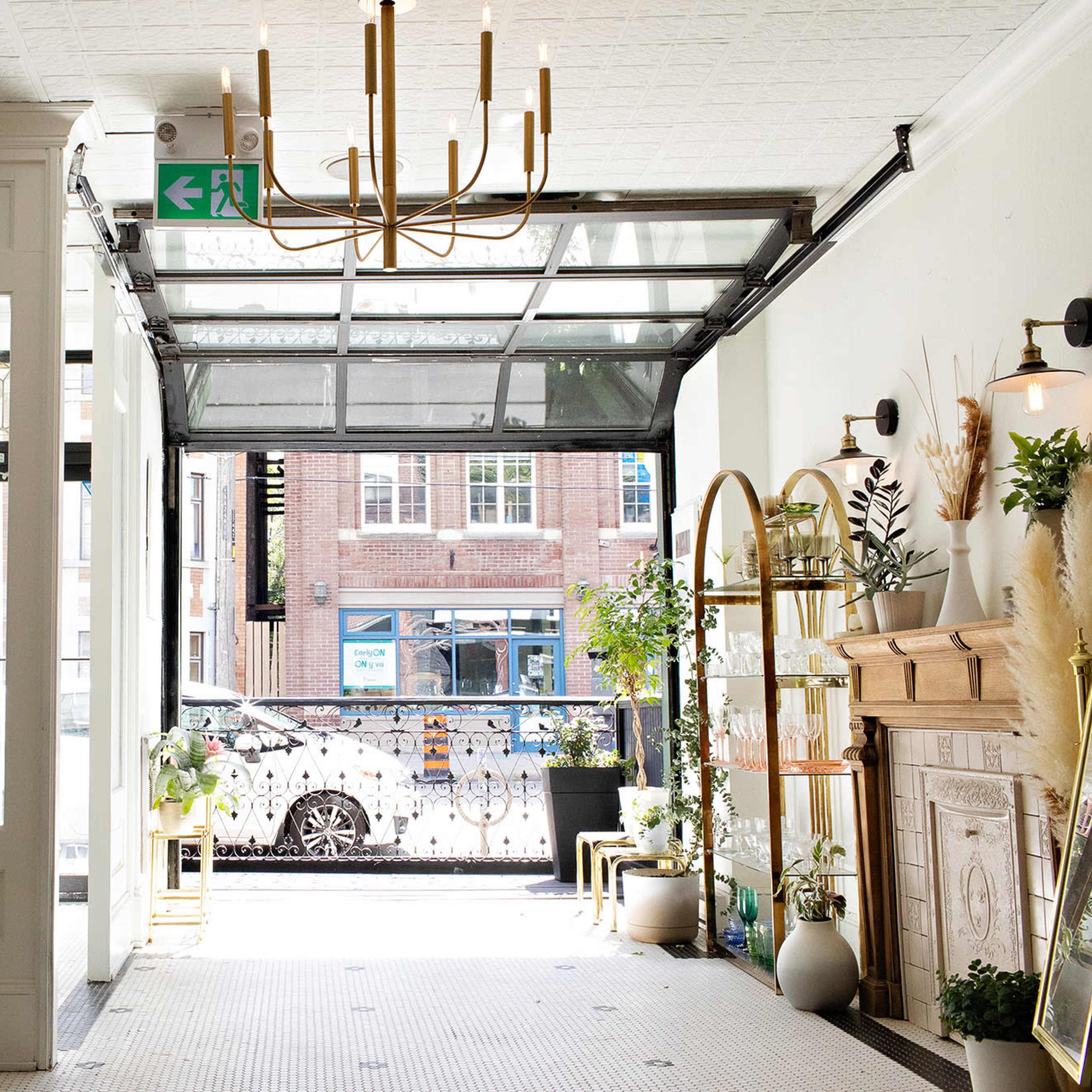 A cafe interior features a large open garage door, allowing natural light to illuminate plants and decor while providing a view of the street outside.