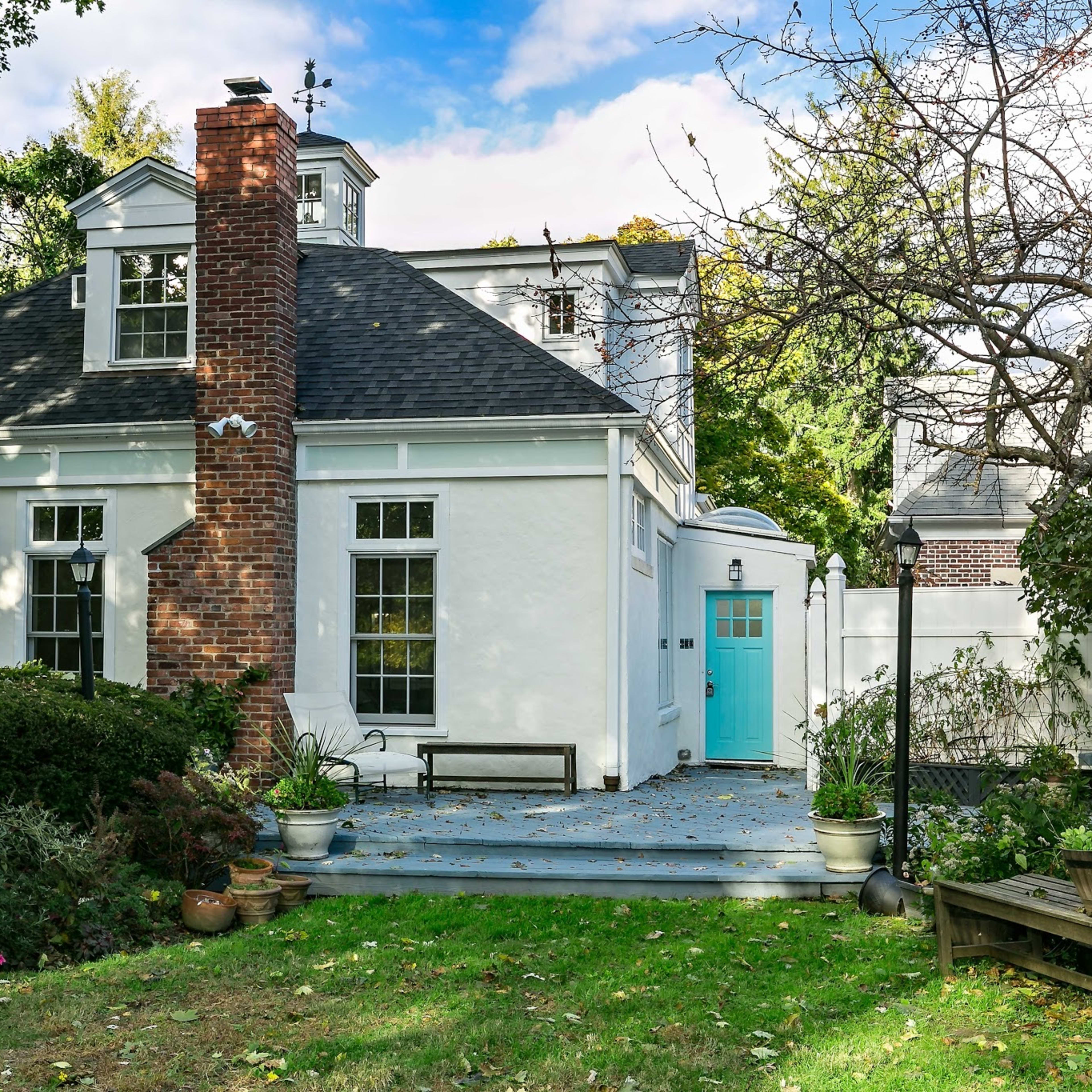 A white house with a brick chimney features a blue door and is surrounded by a well-maintained yard with trees and shrubs.