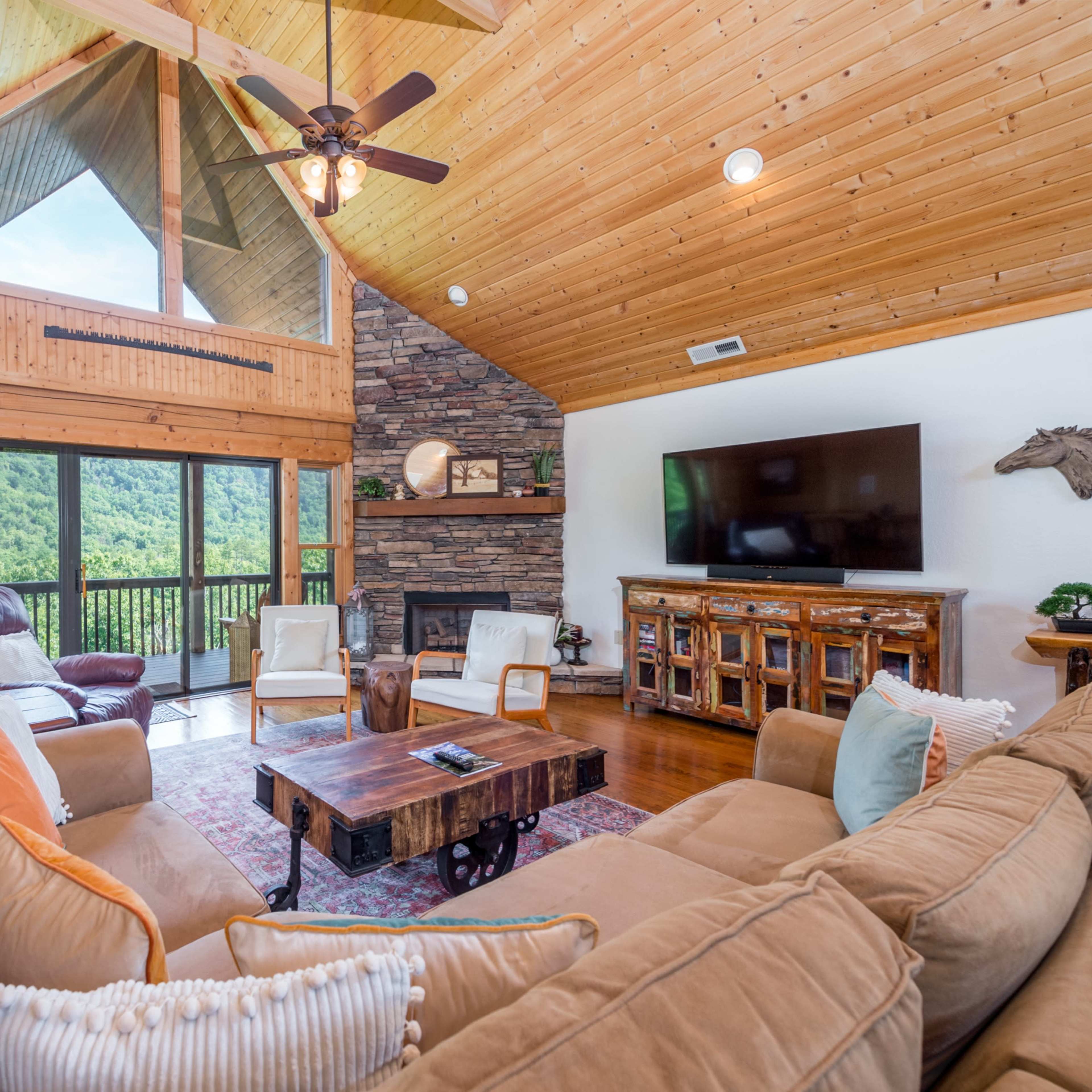 A spacious living room features a large sectional sofa, wooden ceiling beams, a stone fireplace, and a view of lush greenery through large windows.