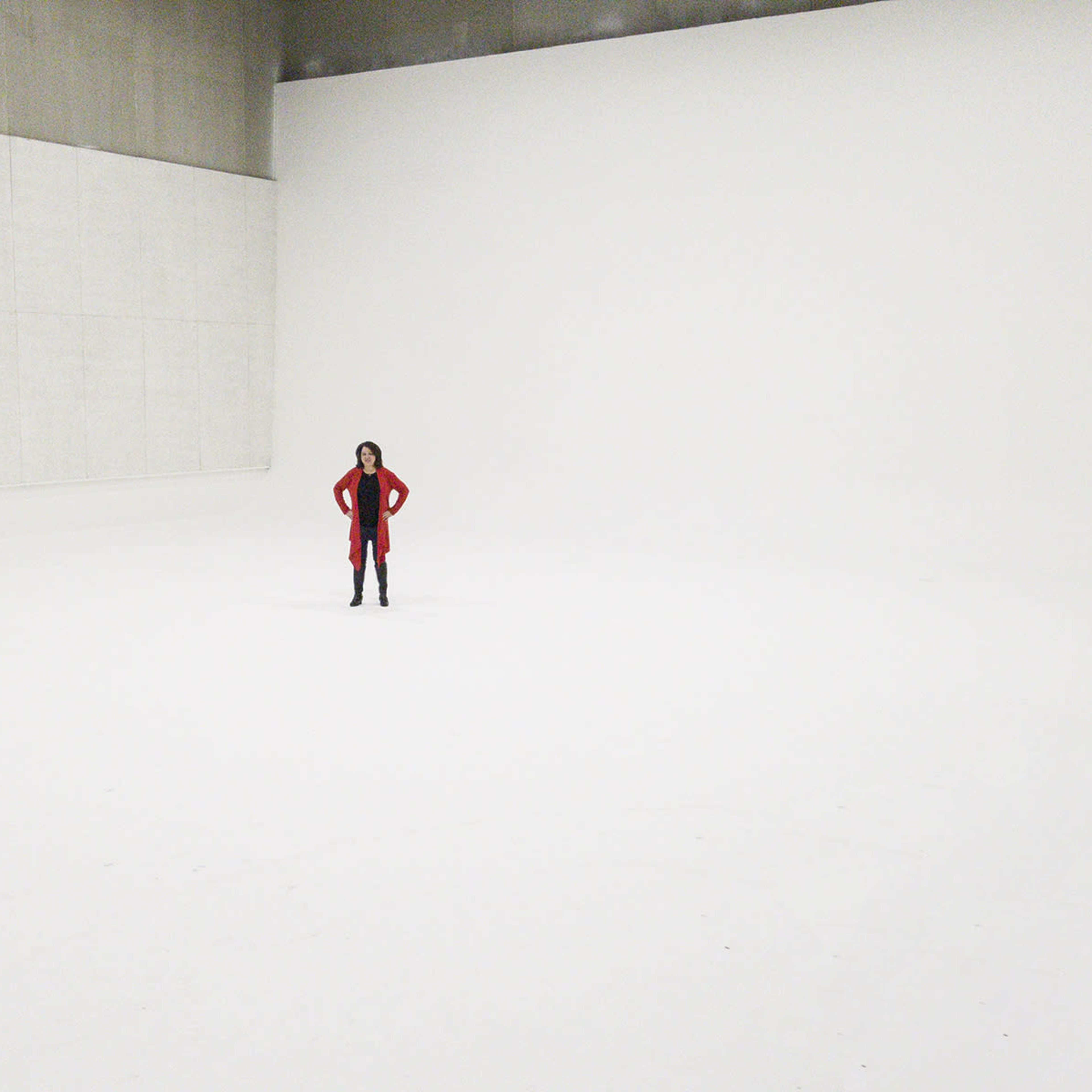 A person stands alone in a large, empty, white room with a smooth floor and bare walls.
