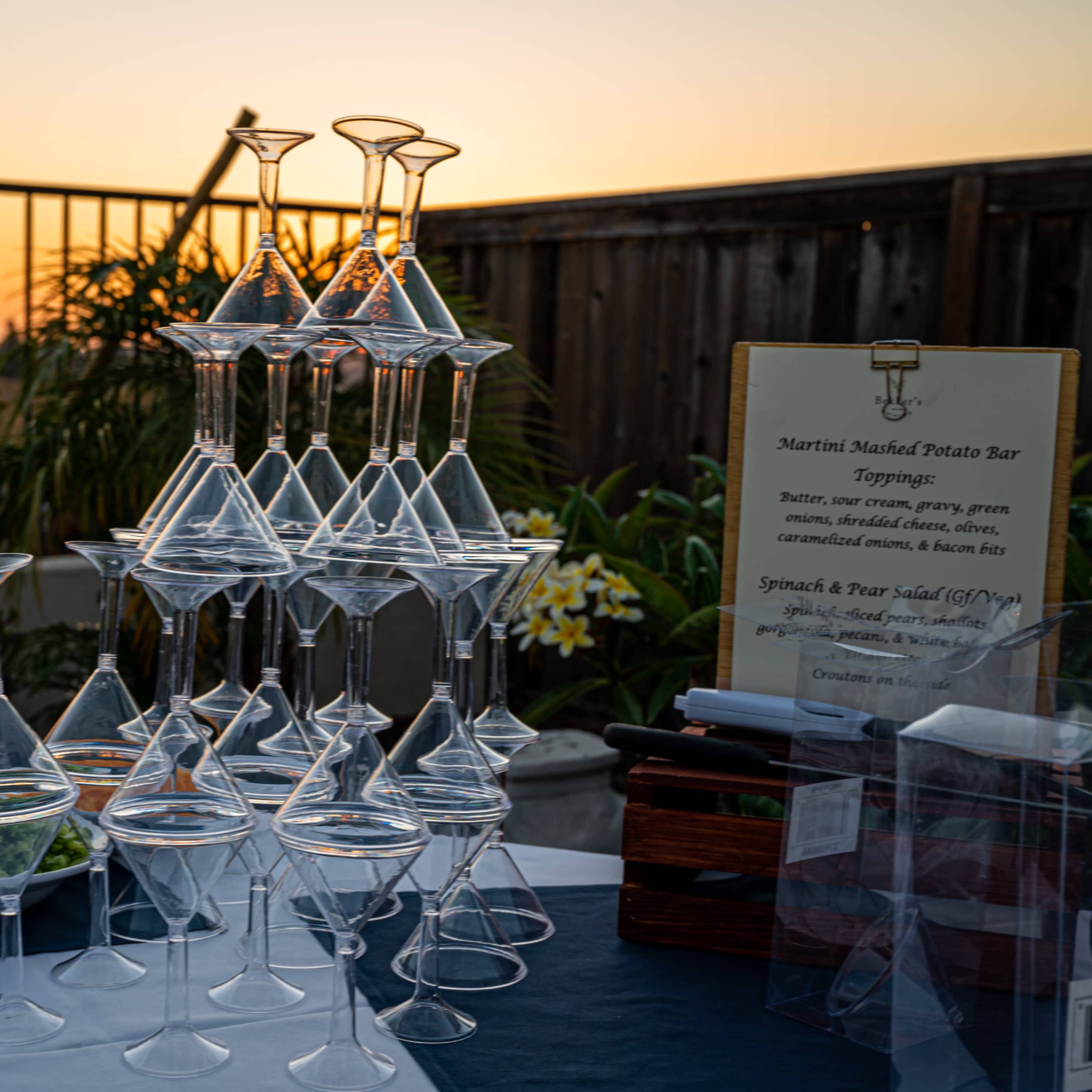 A stack of clear martini glasses is arranged on a table beside a sign for a potato bar, with a sunset in the background.